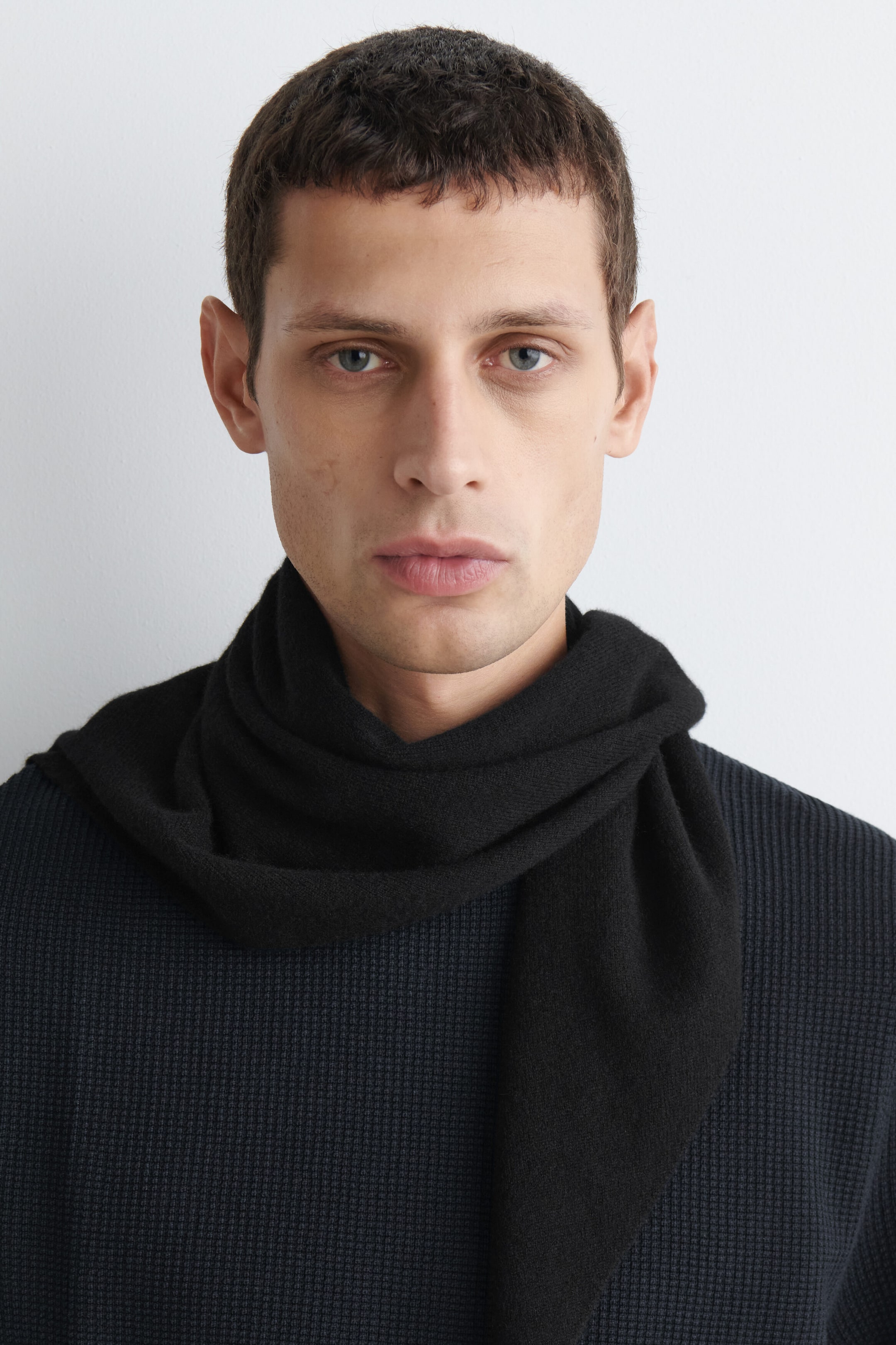 A black cashmere triangular scarf is draping loosely around the neck in a front-facing view, with one pointed end hanging down the chest over a dark, textured waffle-knit top. A black cashmere triangular scarf is draping loosely around the neck in a front-facing view, with one pointed end hanging down the chest over a dark, textured waffle-knit top.