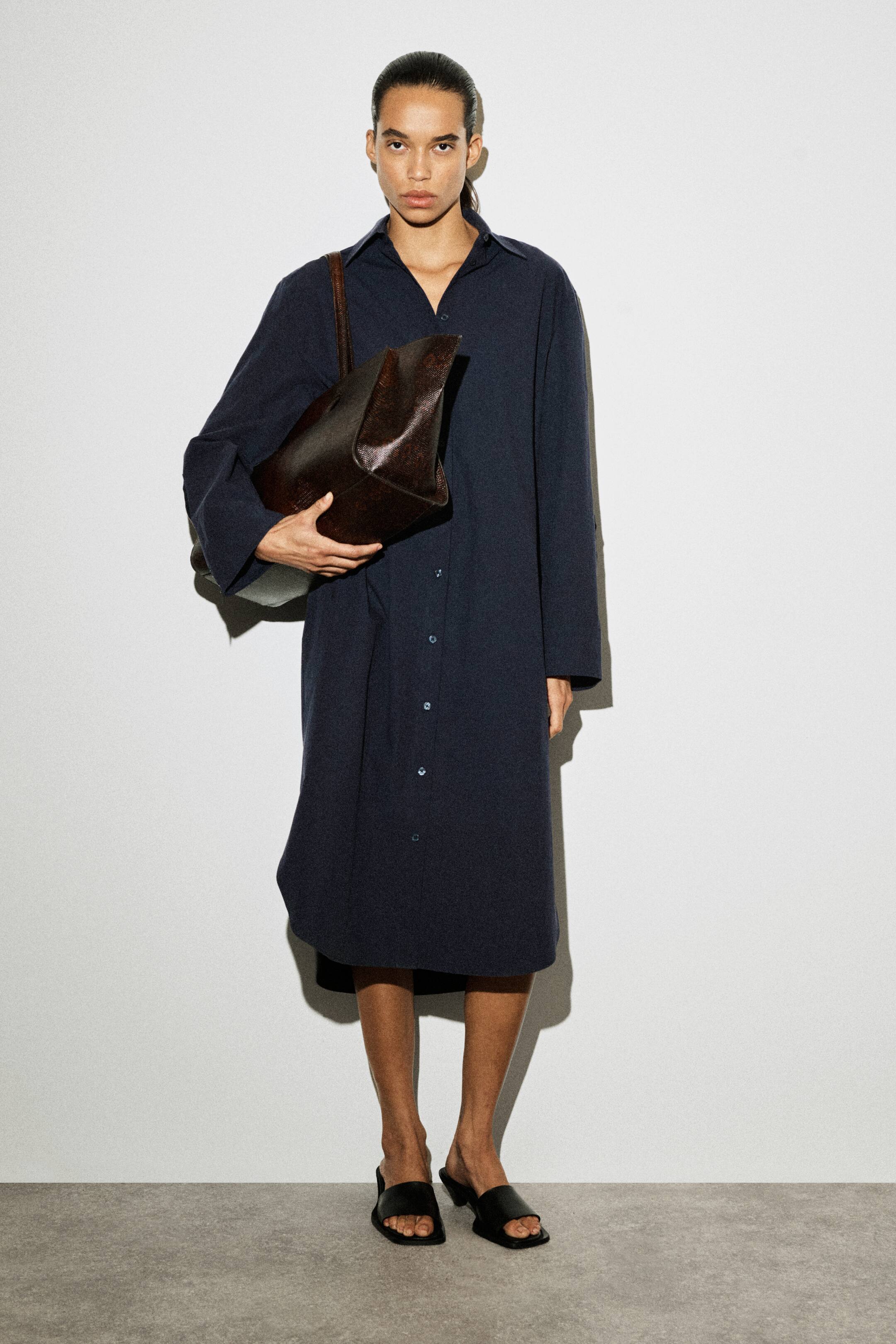 Grösseres Bild ansehen: Ein marineblaues, strukturiertes Hemdkleid mit langen Ärmeln wird von vorne leicht schräg präsentiert, wodurch der lockere Schnitt hervorgehoben wird. Es verfügt über einen spitzen Kragen, eine durchgehende Knopfleiste mit hellen Knöpfen und einen abgerundeten Saum, der bis zur Mitte der Wade fällt.