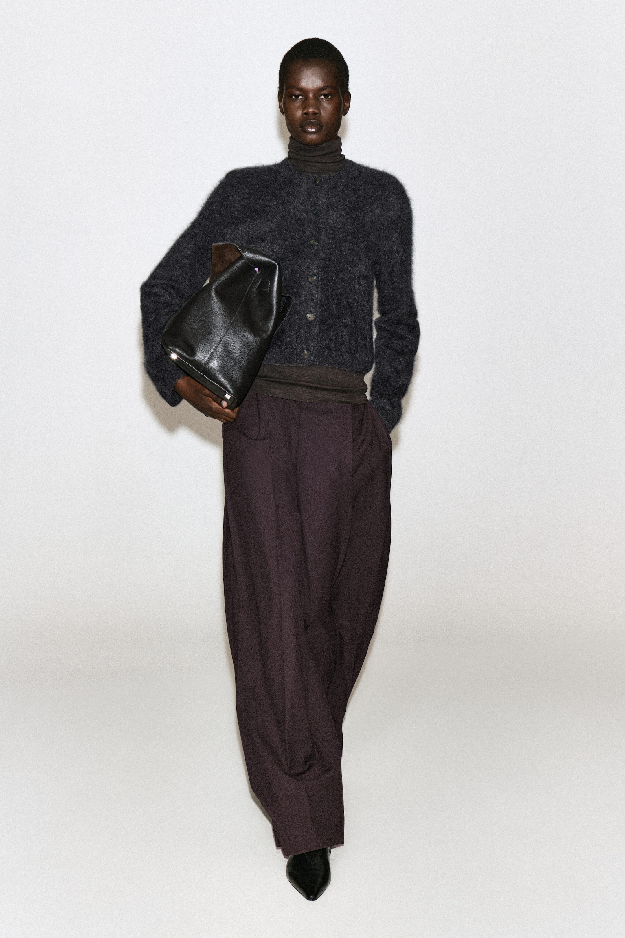 The brushed pure cashmere crew-neck cardigan in charcoal grey is worn buttoned up by the model, showcasing its soft, textured surface. It is layered over a dark brown turtleneck top and styled with dark wide-leg trousers, while the model is holding a black leather bag. The brushed pure cashmere crew-neck cardigan in charcoal grey is worn buttoned up by the model, showcasing its soft, textured surface. It is layered over a dark brown turtleneck top and styled with dark wide-leg trousers, while the model is holding a black leather bag.
