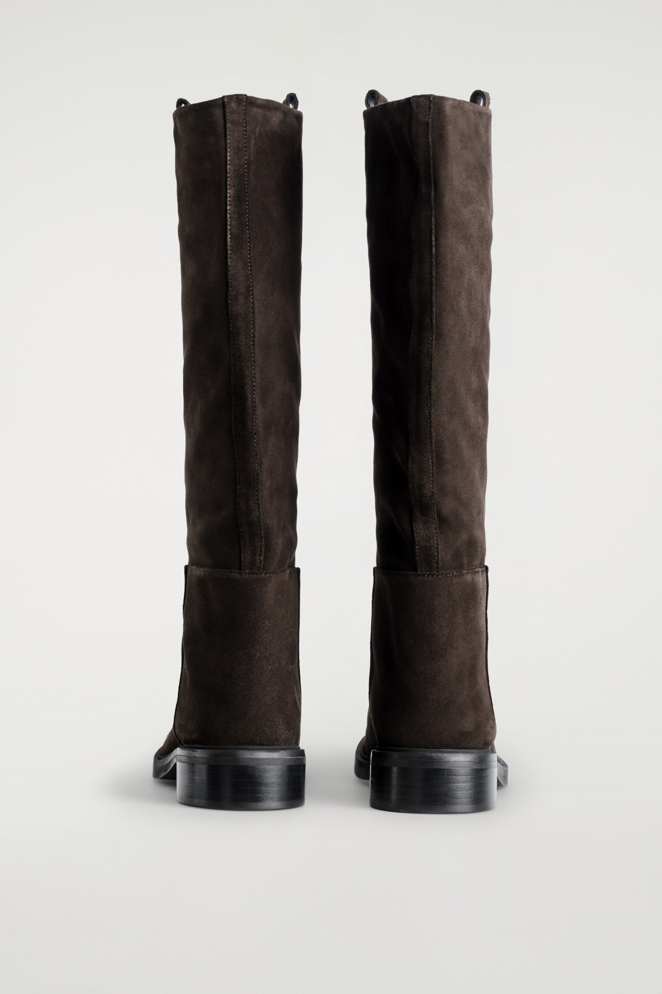 Größeres Bild ansehen: Zwei hohe, dunkelbraune Wildlederstiefel stehen, von hinten präsentiert. Jeder Stiefel verfügt über einen kniehohen Schaft mit einer zentralen vertikalen Naht und tonalen Zuglaschen oben, die auf einem niedrigen, gestapelten Blockabsatz ruhen.
