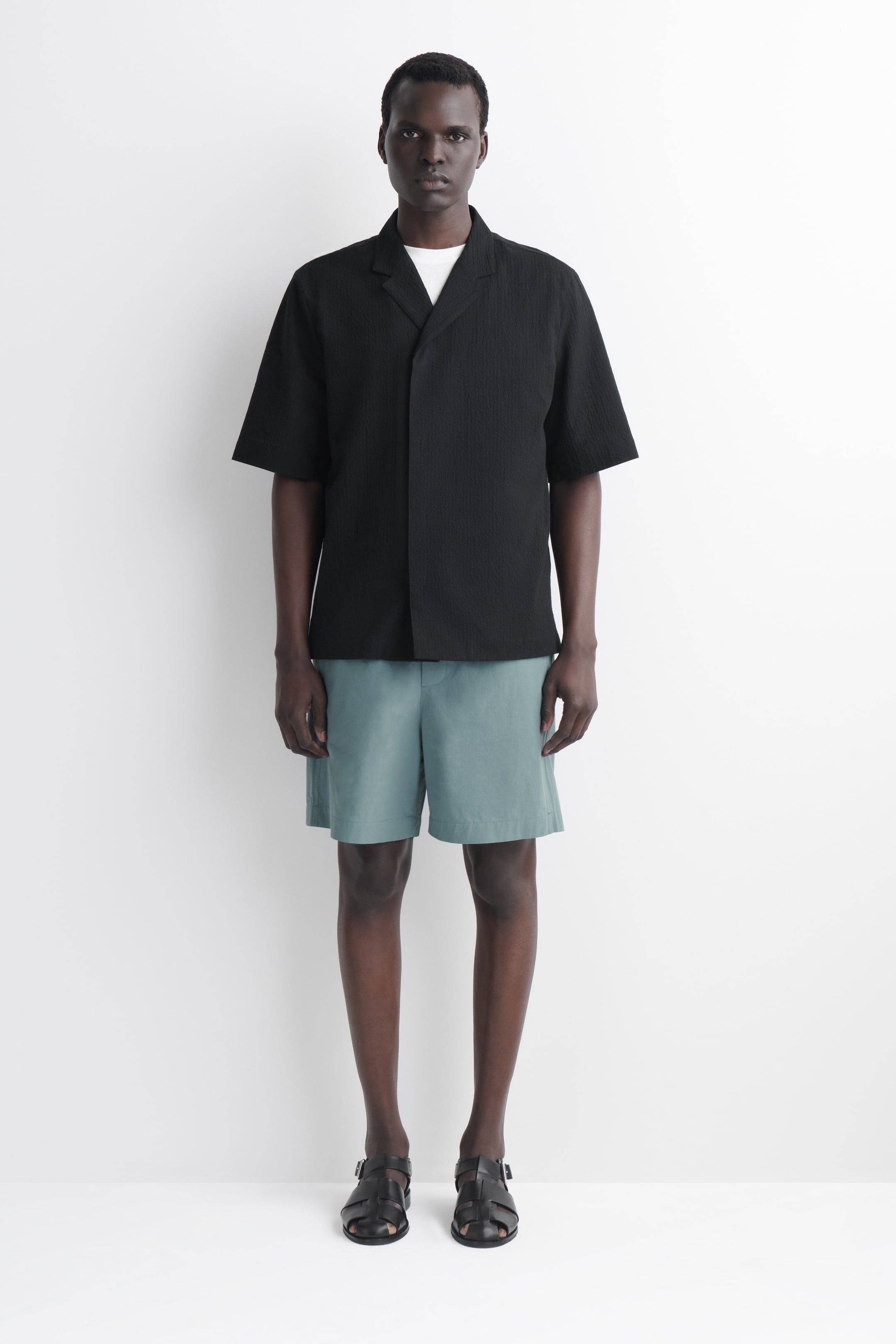 Visualizza immagine più grande: Una camicia nera a maniche corte presenta un colletto a revers e un tessuto strutturato. Viene indossata aperta sopra una t-shirt bianca, rivelando la sua vestibilità rilassata.
