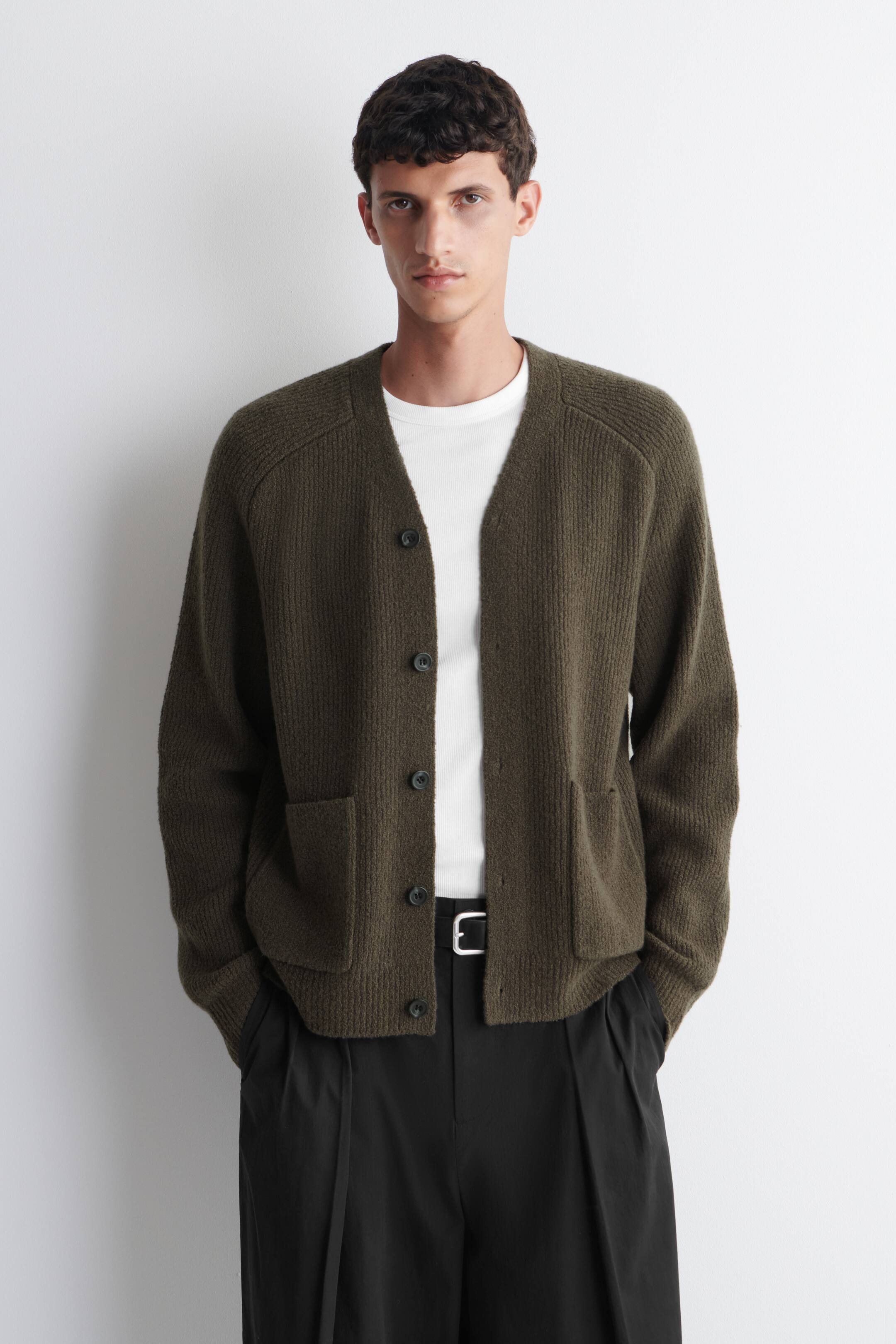 Agrandir l'image: Un cardigan en maille côtelée vert olive, avec un col en V, une fermeture à boutons et deux poches plaquées à l'avant, est porté ouvert par le mannequin sur un t-shirt blanc.