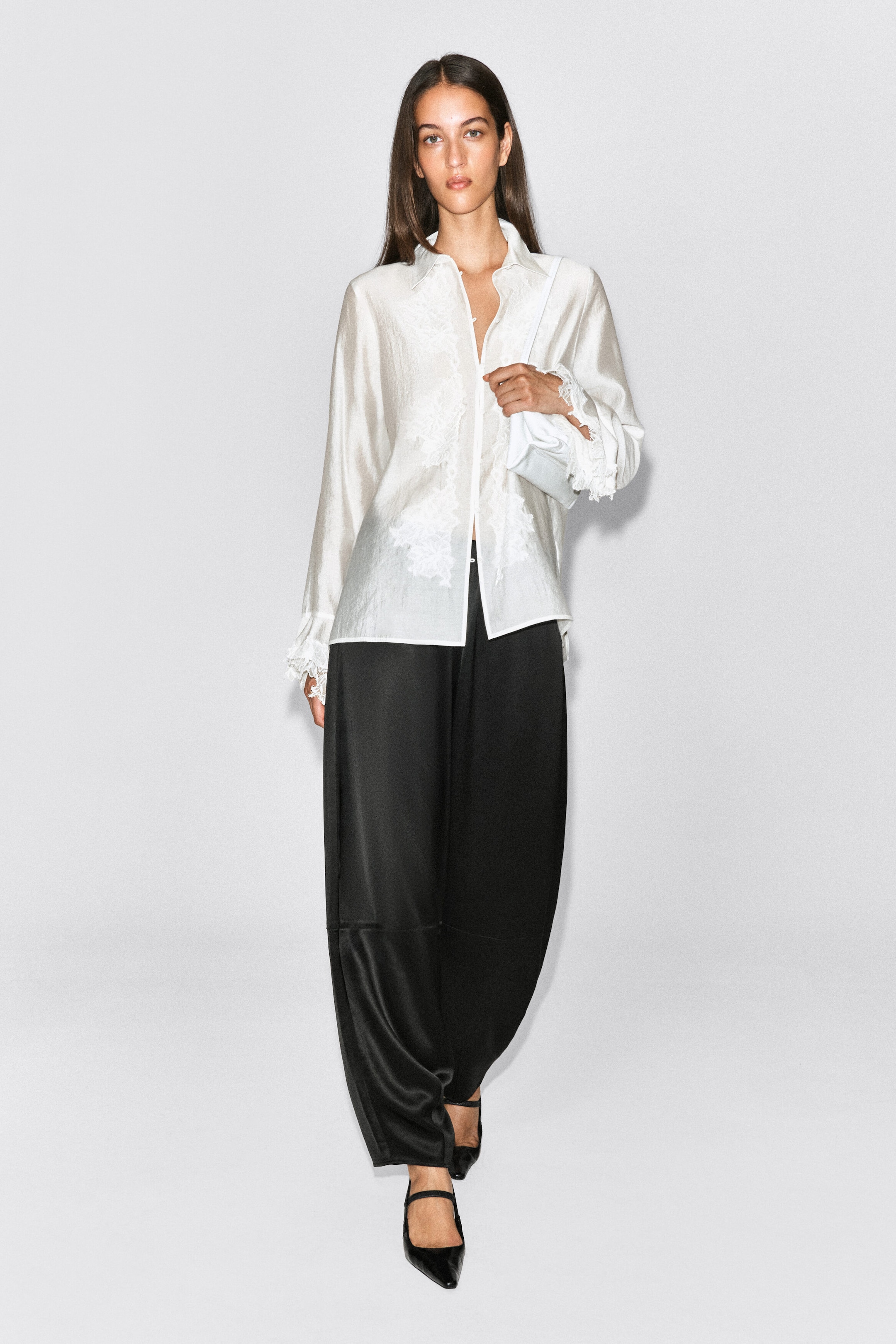 These black satin trousers, featuring a curved barrel-leg silhouette, are worn by the model. The trousers are styled with a white long-sleeve satin shirt and black pointed-toe heels. These black satin trousers, featuring a curved barrel-leg silhouette, are worn by the model. The trousers are styled with a white long-sleeve satin shirt and black pointed-toe heels.