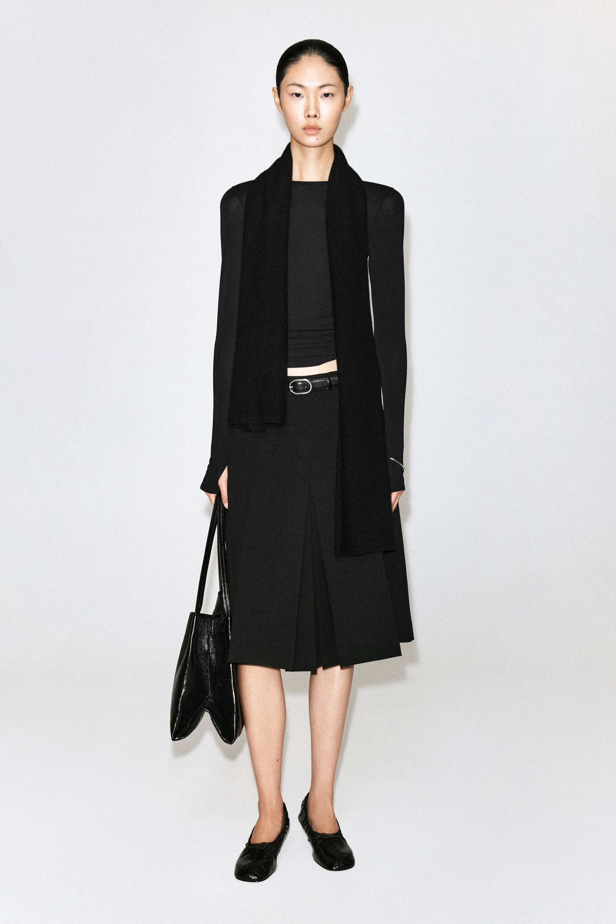 Une jupe plissée noire mi-longue, arrivée au genou, avec une fine taille ceinturée est portée par le mannequin, associée à un haut ajusté à manches longues et à une écharpe noire drapée. Un cabas en cuir texturé noir est tenu à la main par le mannequin, complétant la tenue monochrome.