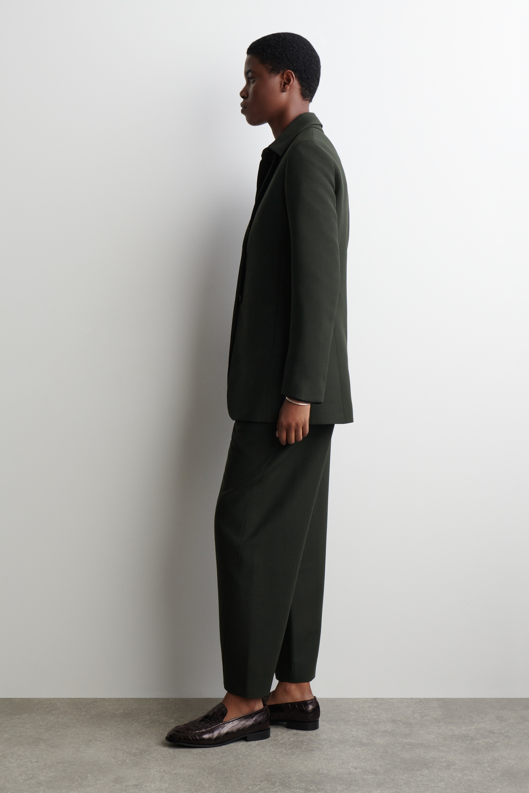 Grösseres Bild ansehen: Ein dunkelgrüner Blazer und passende weite Hosen werden aus einer Seitenansicht präsentiert. Der Blazer zeichnet sich durch eine entspannte Passform mit einem einfachen Kragen und langen Ärmeln aus, und die langen Hosen fallen leicht über dunkle, strukturierte Loafer.