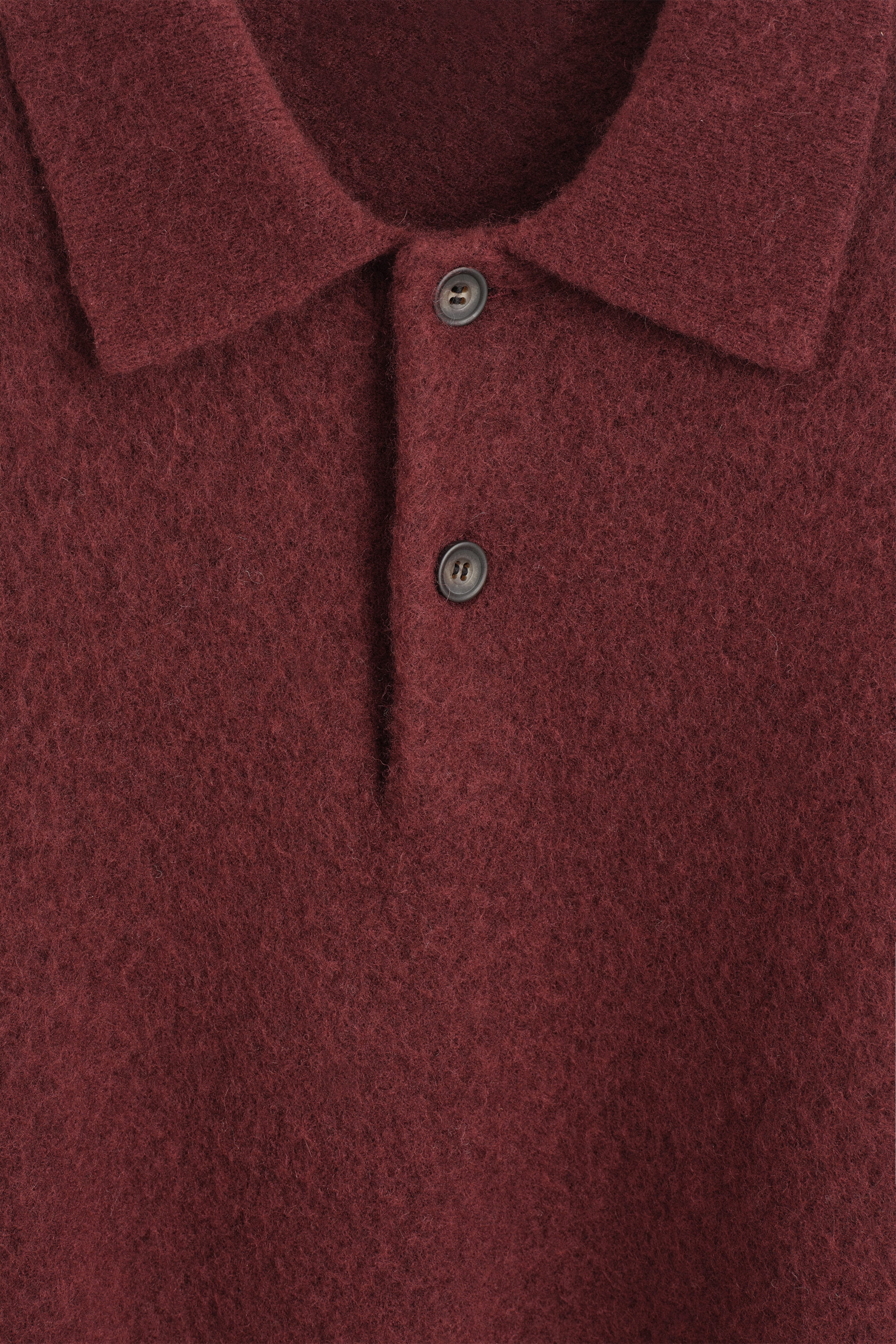 This front close-up of the burgundy brushed wool polo shirt highlights its soft, fuzzy texture. The shirt features a classic collar and a two-button placket with dark-colored buttons. This front close-up of the burgundy brushed wool polo shirt highlights its soft, fuzzy texture. The shirt features a classic collar and a two-button placket with dark-colored buttons.