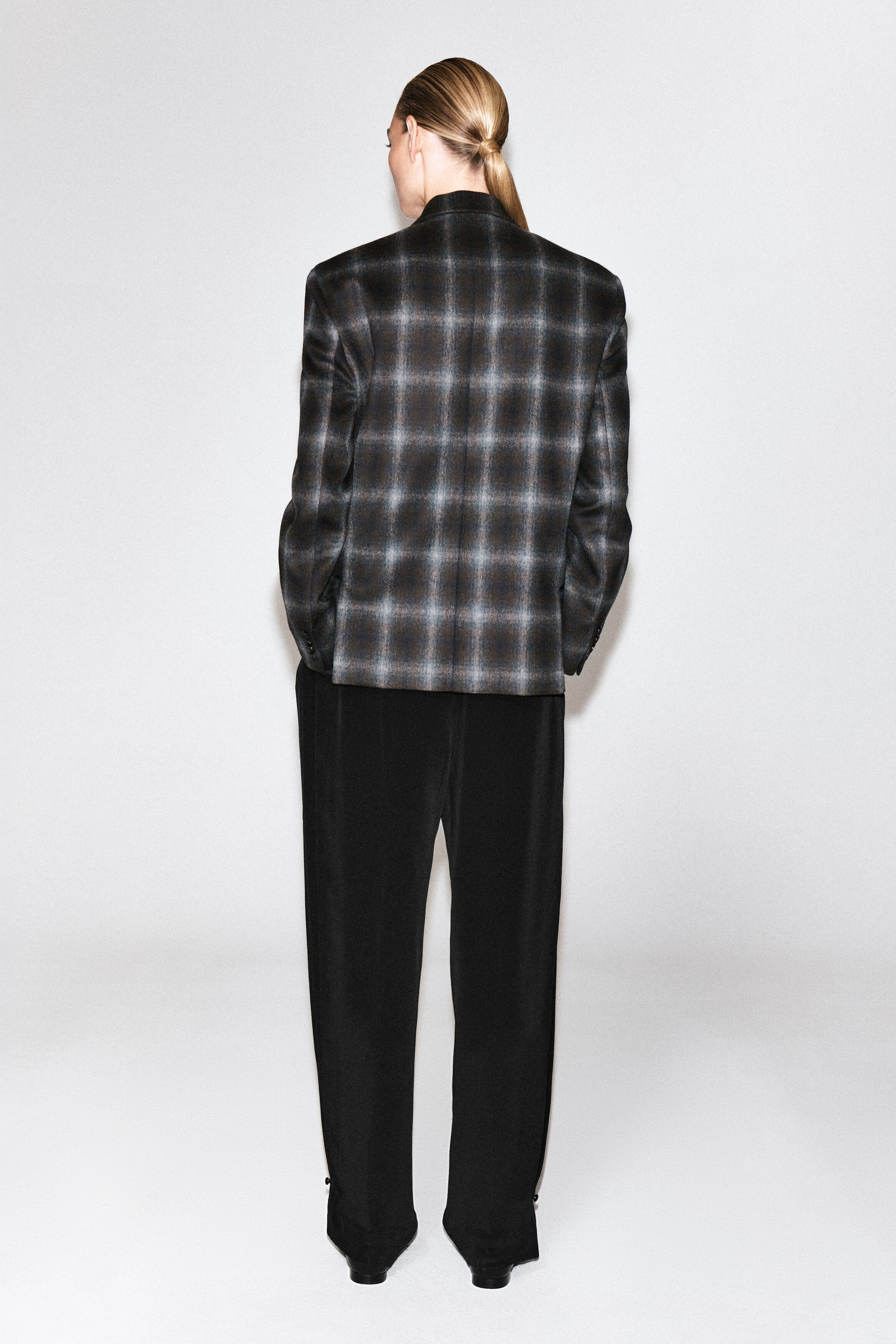 Un blazer en mélange de laine à carreaux, avec un motif tartan sombre dans des tons gris et brun, est porté par le mannequin, qui se tient de dos face à l’appareil photo. Le blazer structuré est associé à un pantalon noir coupe ample.