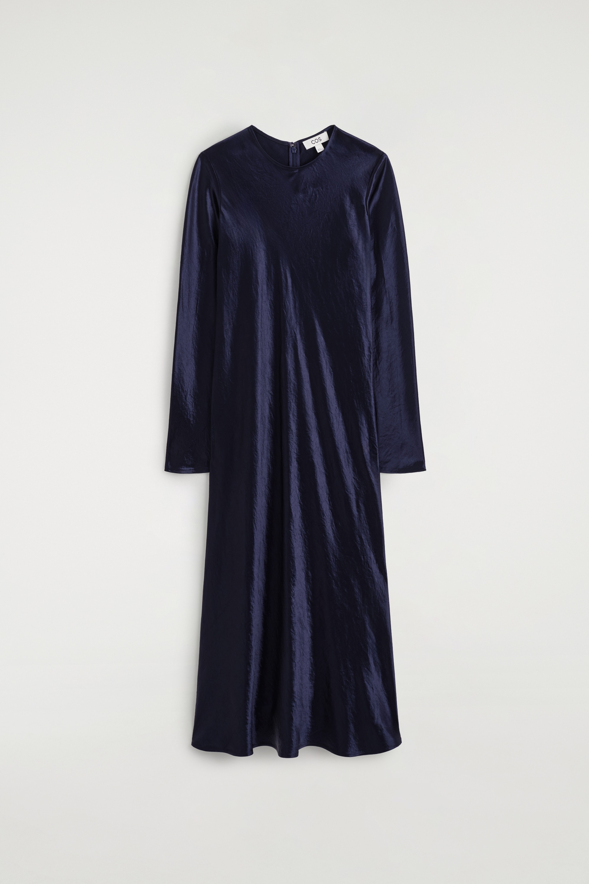 A dark navy blue satin long-sleeved maxi dress with a textured metallic sheen is being worn by the model. The dress features a round neckline and is styled with a black clutch bag tucked under the modelâs arm and black pointed-toe shoes.
