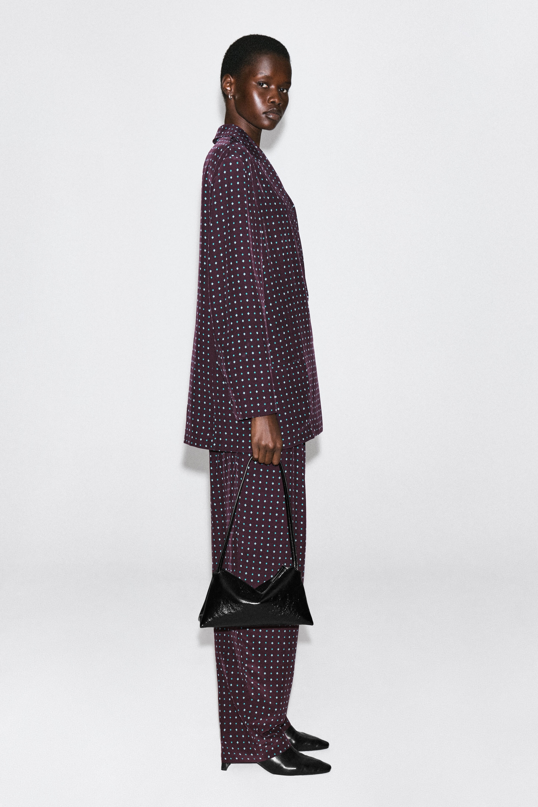 The diamond-print double-breasted blazer, featuring a deep plum fabric with a small teal diamond pattern, is shown facing right to highlight its relaxed fit and structured shoulders. It is being worn with matching trousers, creating a coordinated and fluid silhouette. The diamond-print double-breasted blazer, featuring a deep plum fabric with a small teal diamond pattern, is shown facing right to highlight its relaxed fit and structured shoulders. It is being worn with matching trousers, creating a coordinated and fluid silhouette.