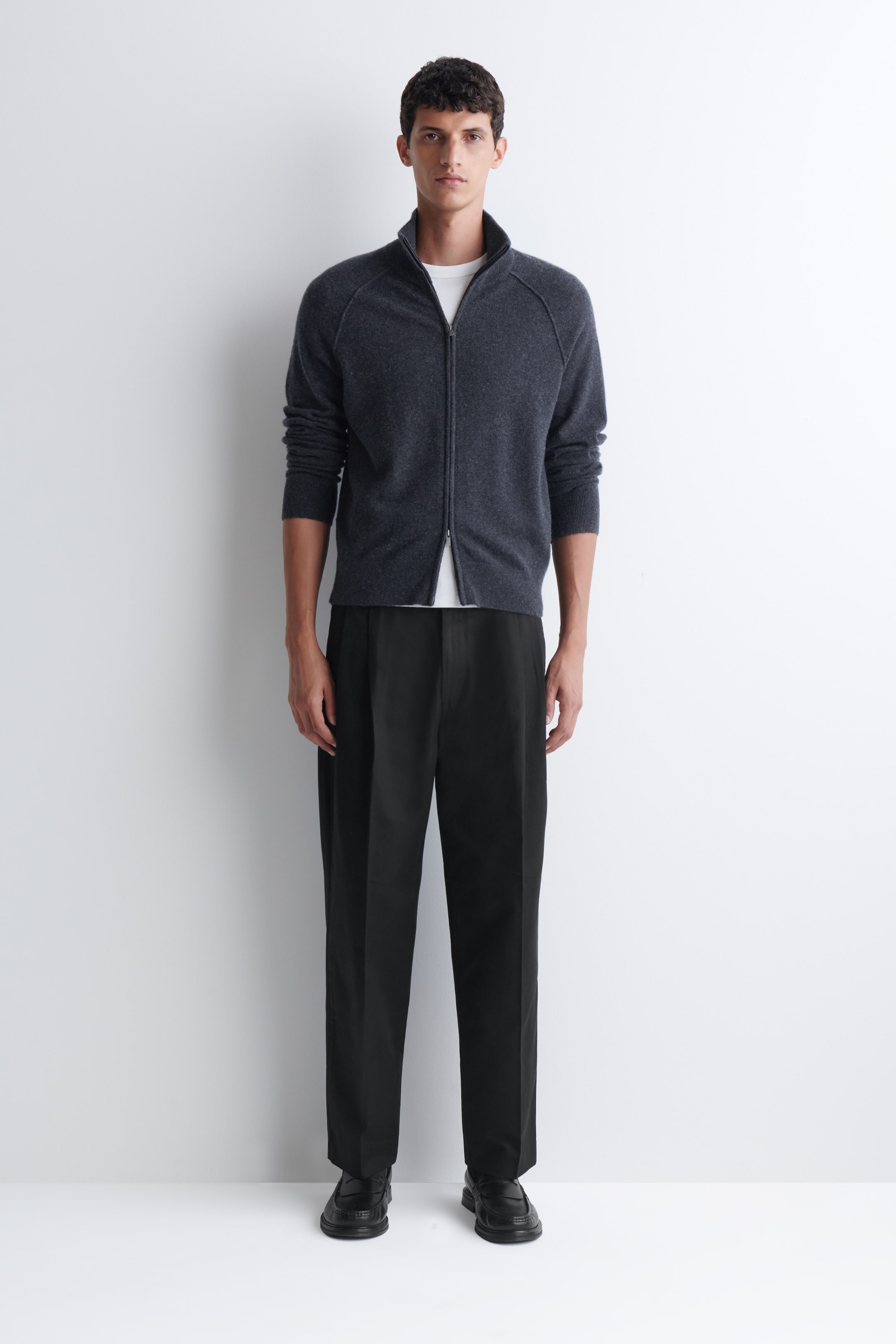 Vezi imaginea mai mare: Pantalonii plisați, cu croială relaxată și tapered, sunt negri, având pliuri în partea din față a taliei și o croială lejeră care se îngustează spre gleznă. Modelul îi poartă, iar aceștia cad puțin deasupra pantofilor negri.