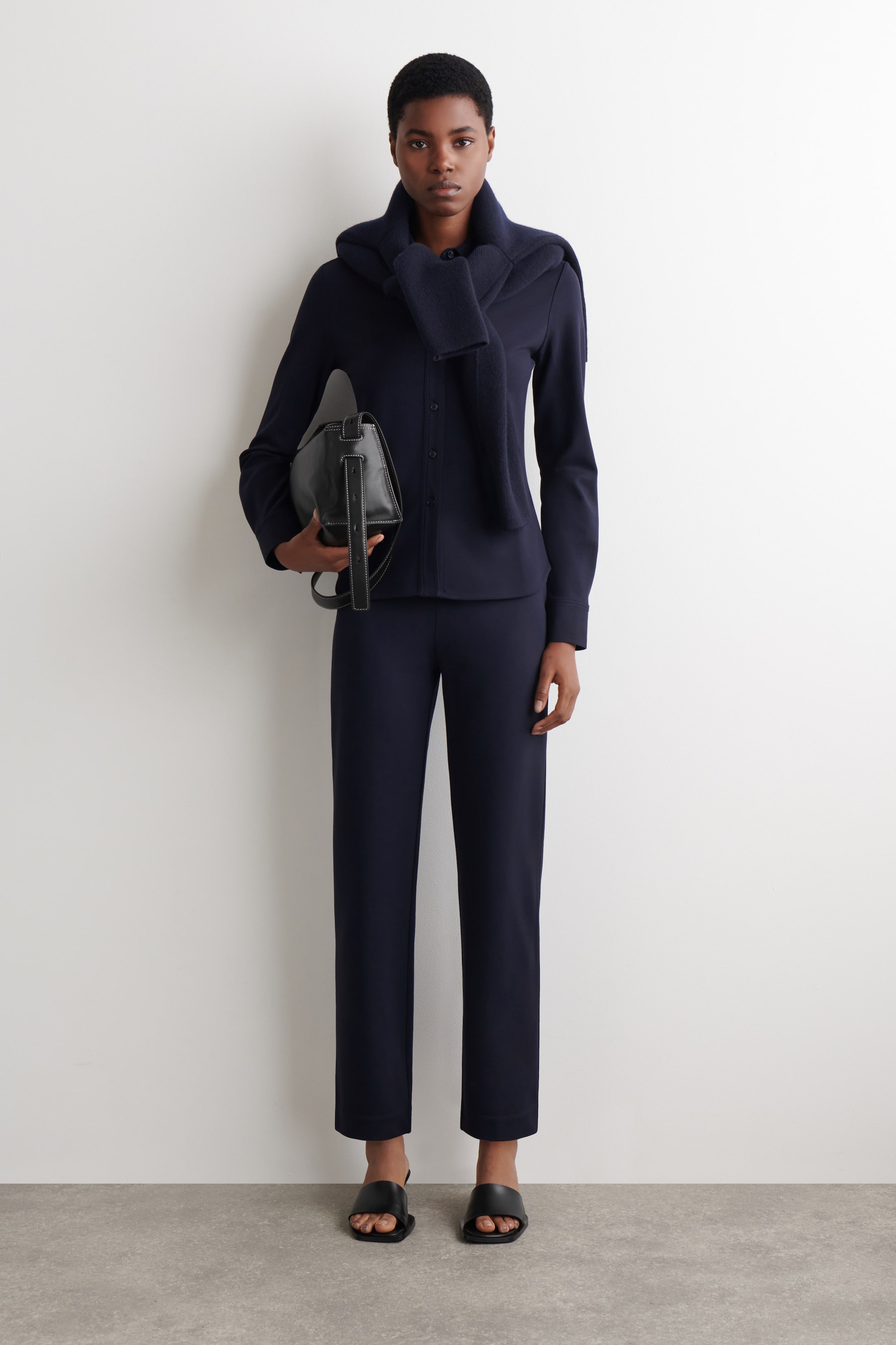 הצגת תמונה גדולה יותר: המוצרים המוצגים הם חולצה כחולה כהה עם שרוולים ארוכים וכפתורים מקדימה ומכנסיים תואמים בגזרה ישרה, עם סוודר כחול כהה מונח על הכתפיים. תיק יד שחור עם תפירה לבנה בקונטרסט מוחזק ביד, ונעלי סנדלים שחורות עם קצה מרובע ננעלות, כל המוצרים מופנים קדימה.