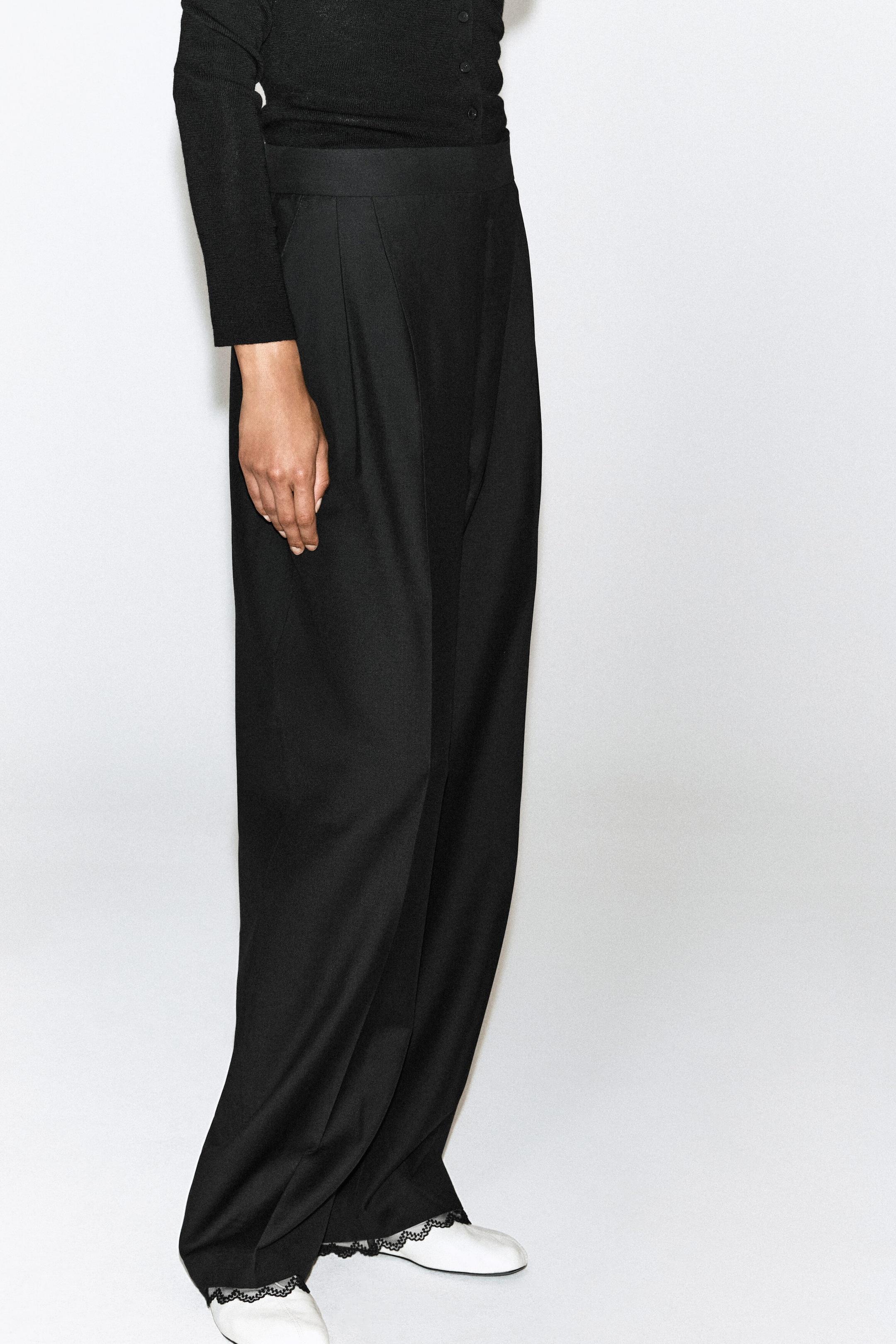 These black high-waisted, wide-leg trousers face right, featuring a structured waistband and front pleats that create a draped silhouette. The trousers fall loosely to the floor and are styled with a tucked-in black long-sleeve top and white shoes. These black high-waisted, wide-leg trousers face right, featuring a structured waistband and front pleats that create a draped silhouette. The trousers fall loosely to the floor and are styled with a tucked-in black long-sleeve top and white shoes.