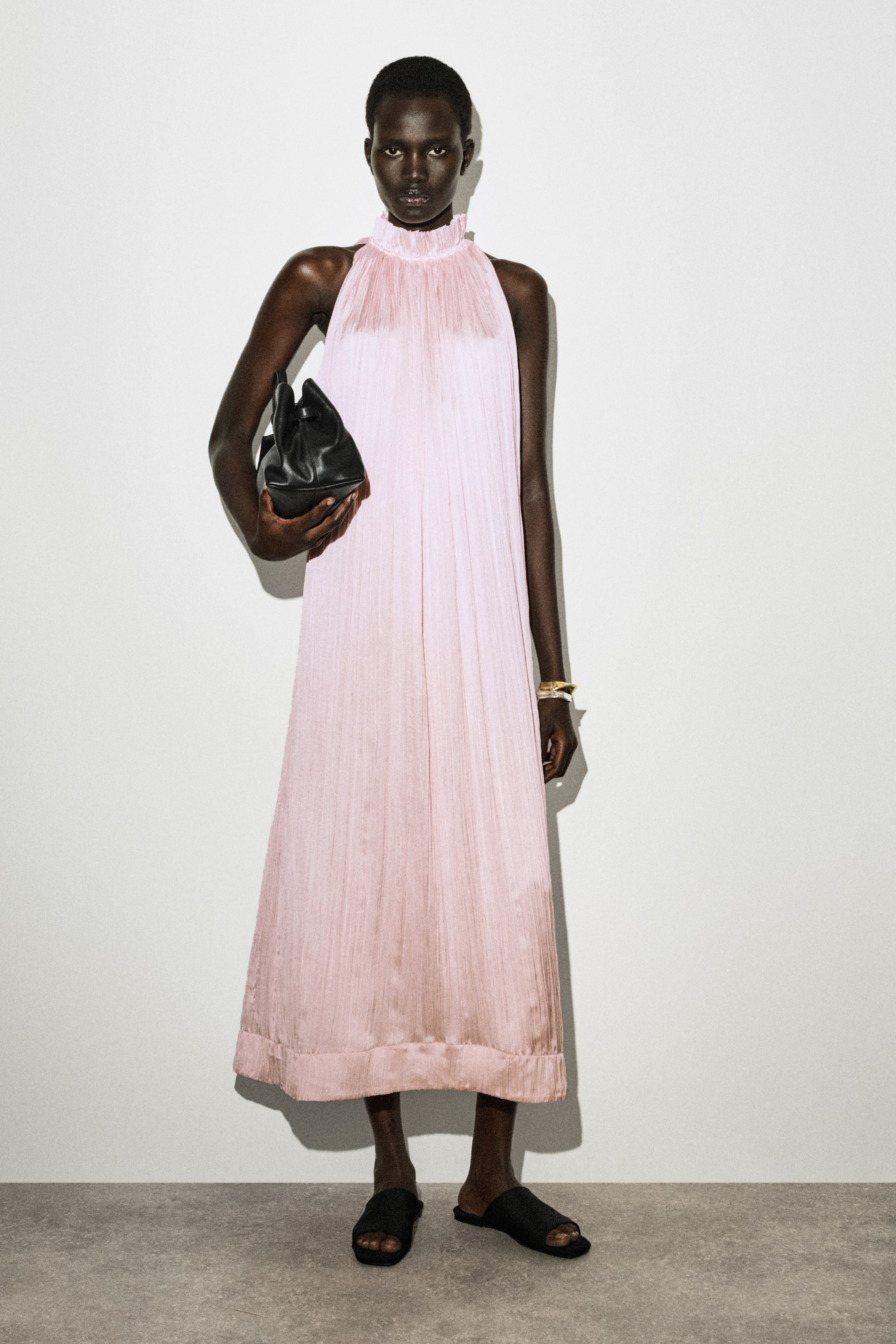 Agrandir l'image: Une robe midi plissée rose clair présente un col montant à volants, des plis verticaux sur toute la longueur et une silhouette fluide, présentée de face. Elle est assortie d'un sac à main noir, de bracelets dorés et de sandales noires à enfiler.