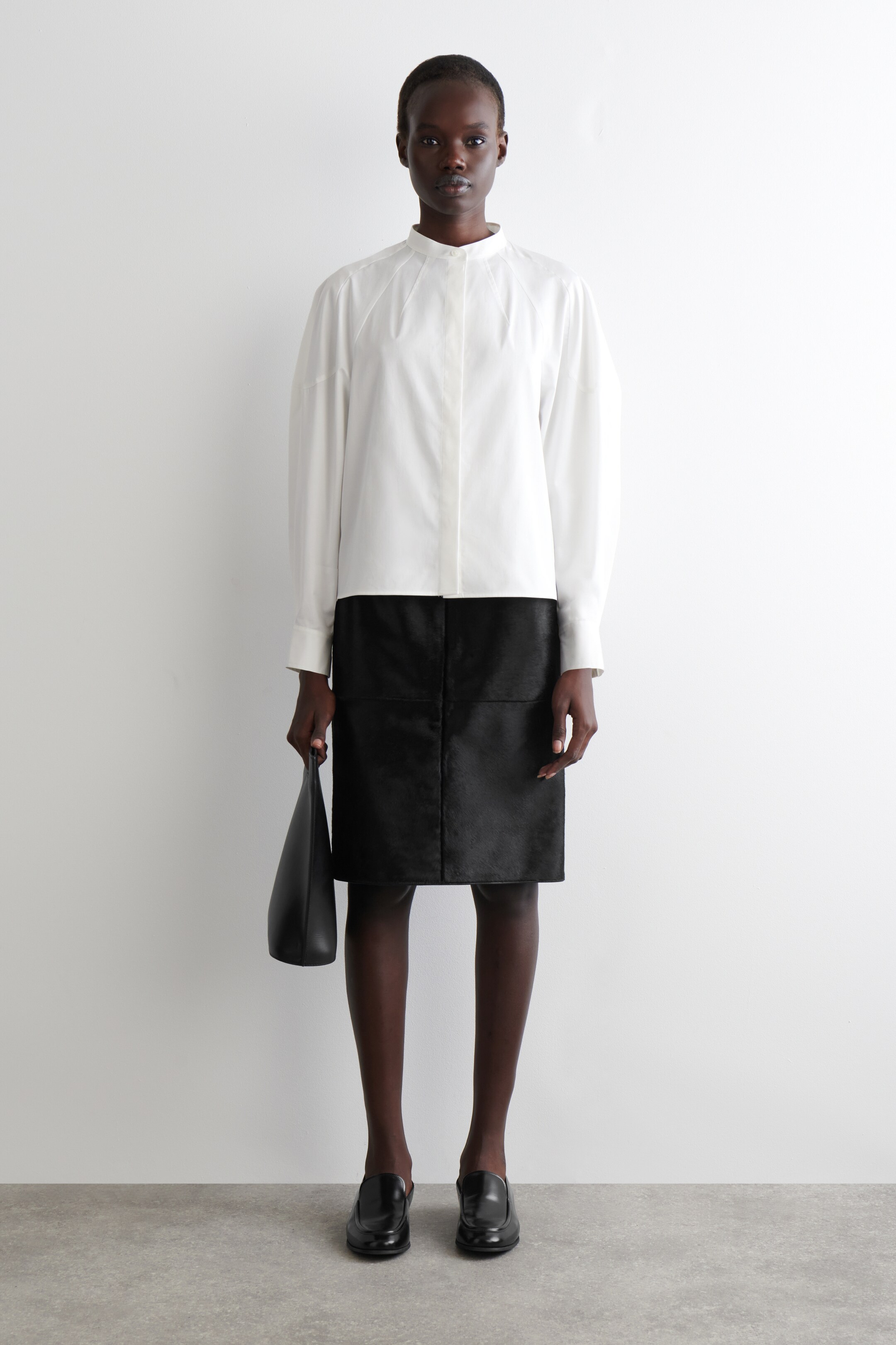 View larger image: A white long-sleeved shirt with a band collar and hidden placket is being worn, featuring a relaxed fit and dropped shoulders. It is styled with a black knee-length skirt, detailed with visible paneling, and black classic loafers are being worn. A soft black tote bag is being held, with the full outfit presented from a front-facing angle.