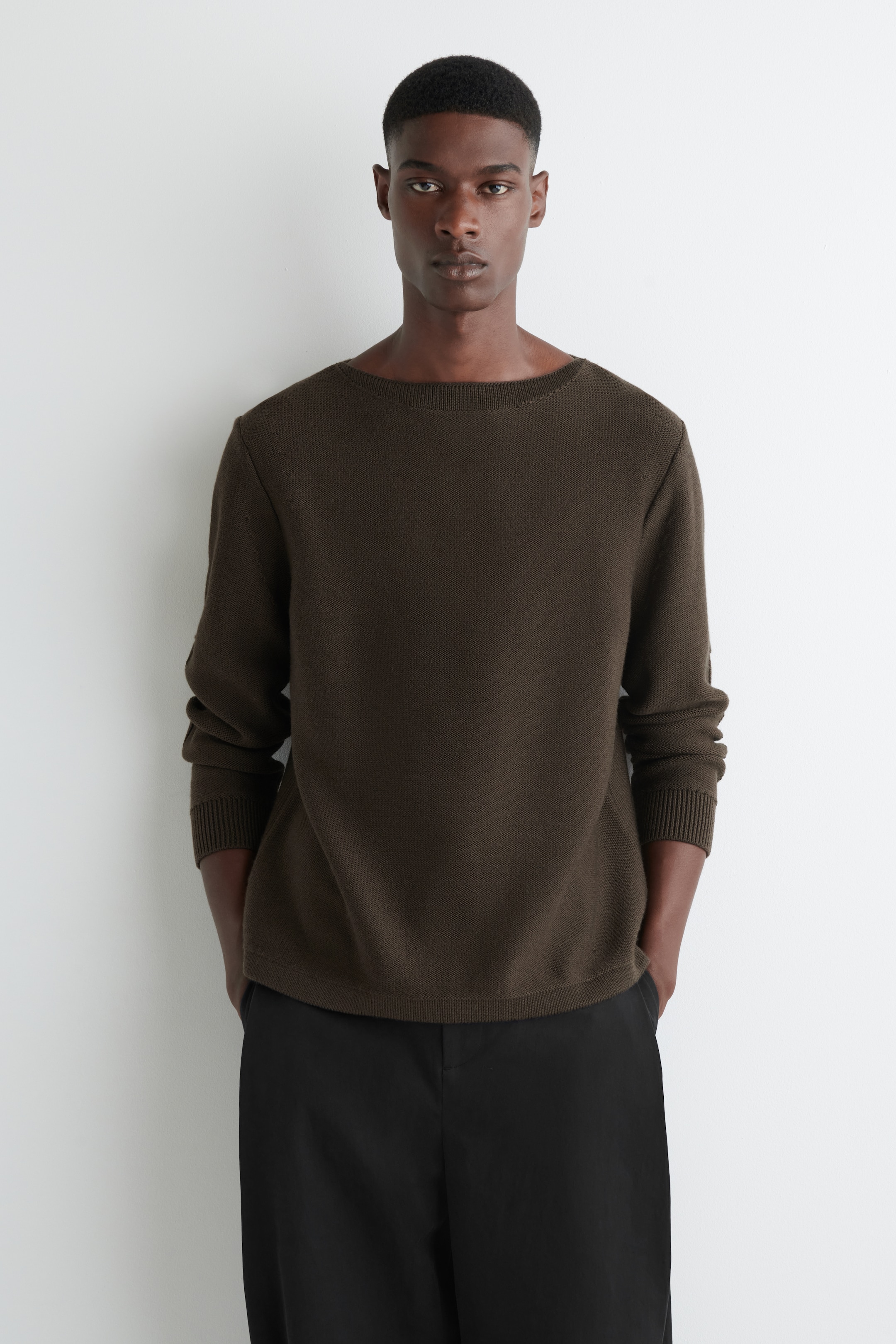 Grösseres Bild ansehen: Ein dunkel olivgrüner Pullover wird frontal präsentiert, mit einem Bootsausschnitt und einer strukturierten Strickart. Er zeigt lange Ärmel mit gerippten Bündchen und einem geraden Saum, getragen über schwarzen Hosen, um seine lockere Passform zu veranschaulichen.