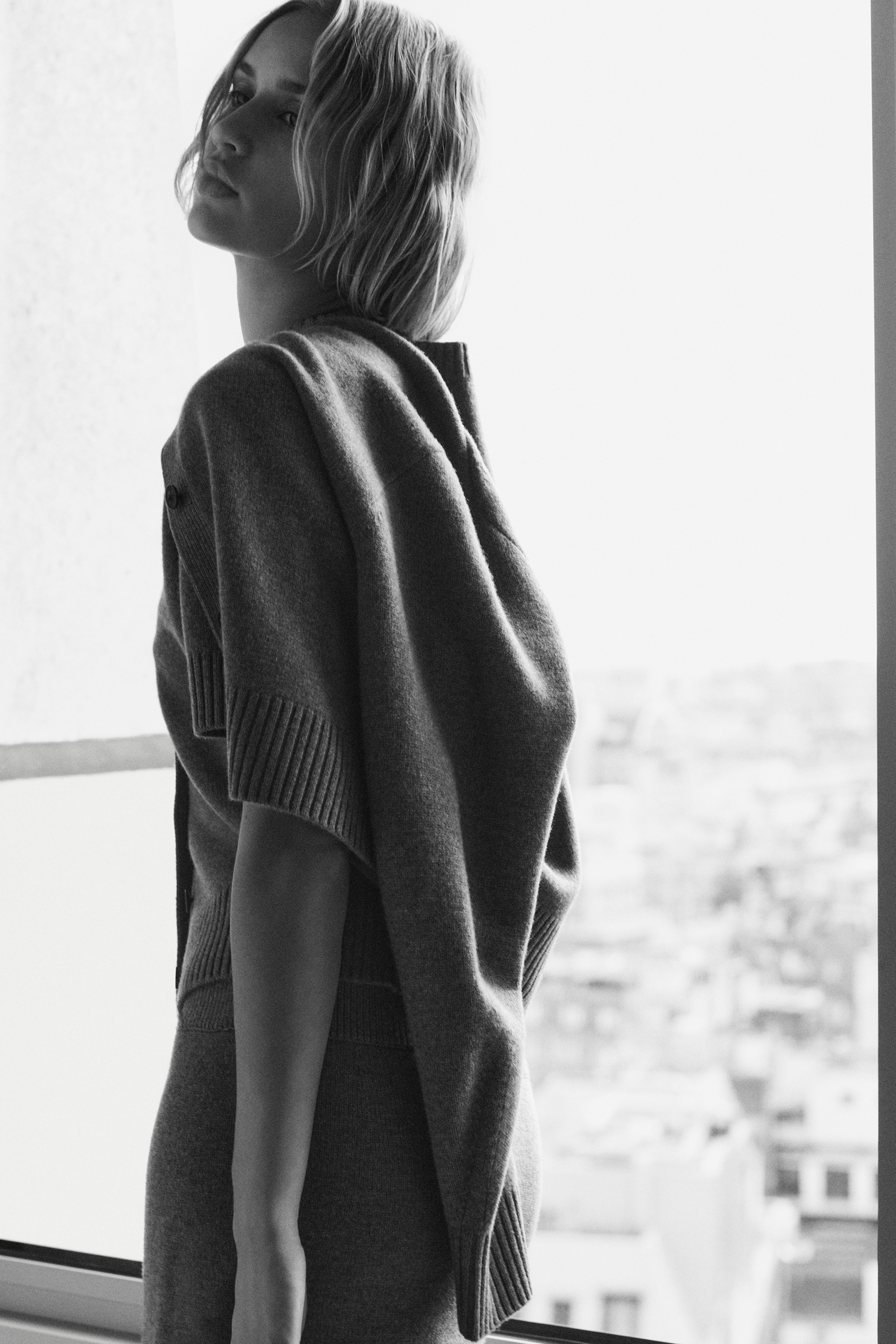 Ingrandisci l'immagine: Un cardigan corto a maniche corte in lana merino color talpa, con polsini a coste e un bottone tono su tono, è drappeggiato sulle spalle. È visibile anche una mini gonna abbinata in lana merino color talpa con bordi a coste.