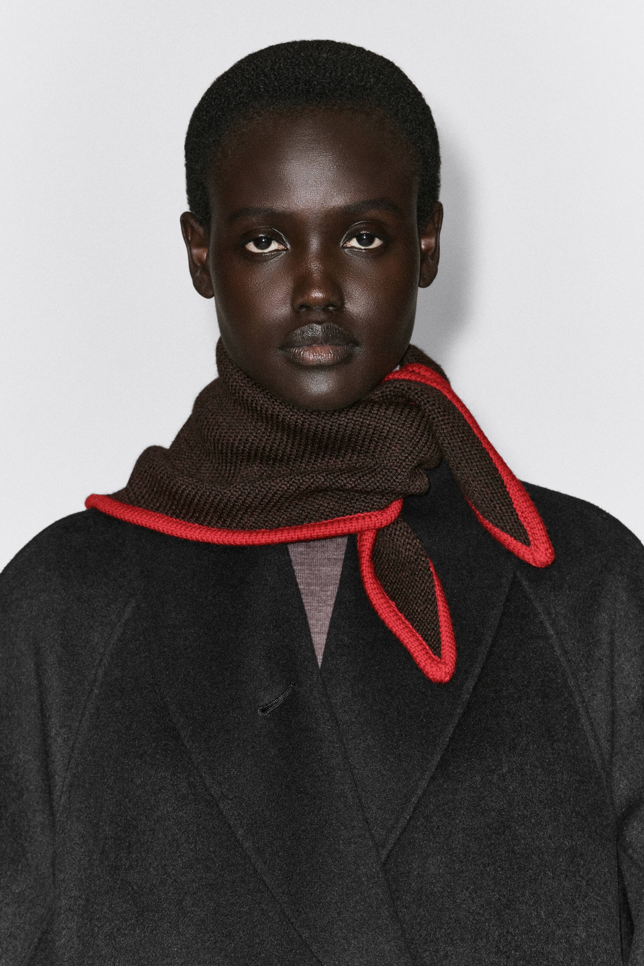 Agrandir l'image: L’écharpe triangle en laine mérinos à bouts contrastés est enroulée autour du cou, orientée vers l’avant, et portée par-dessus un manteau foncé. L’écharpe présente un tricot texturé brun foncé avec une bordure contrastante rouge vif le long de ses bords triangulaires.