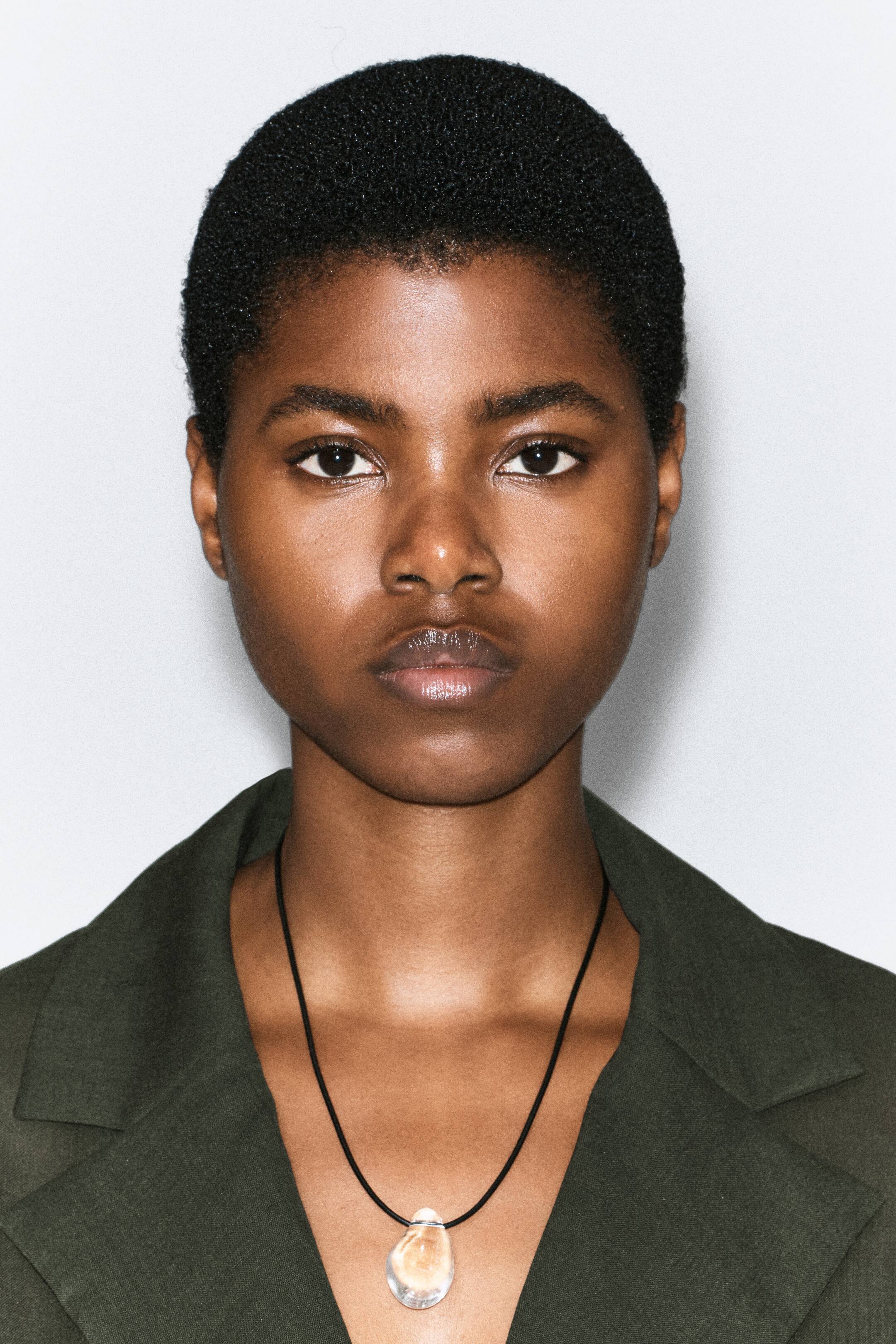Visualizza immagine più grande: Una collana con un cordino nero e un ciondolo trasparente dalla forma organica è indossata dalla modella sopra un blazer verde scuro.