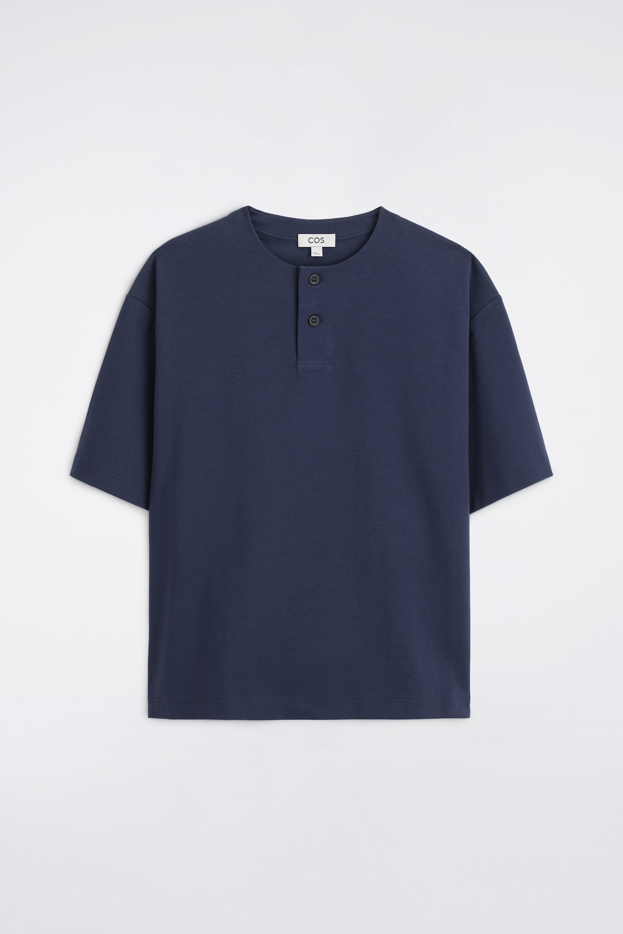 Un t-shirt henley surdimensionné en jersey de coton bleu marine est présenté de face, avec un col rond à patte de boutonnage à deux boutons. Le t-shirt met en valeur une coupe décontractée avec des épaules tombantes, des manches courtes larges et un ourlet droit.