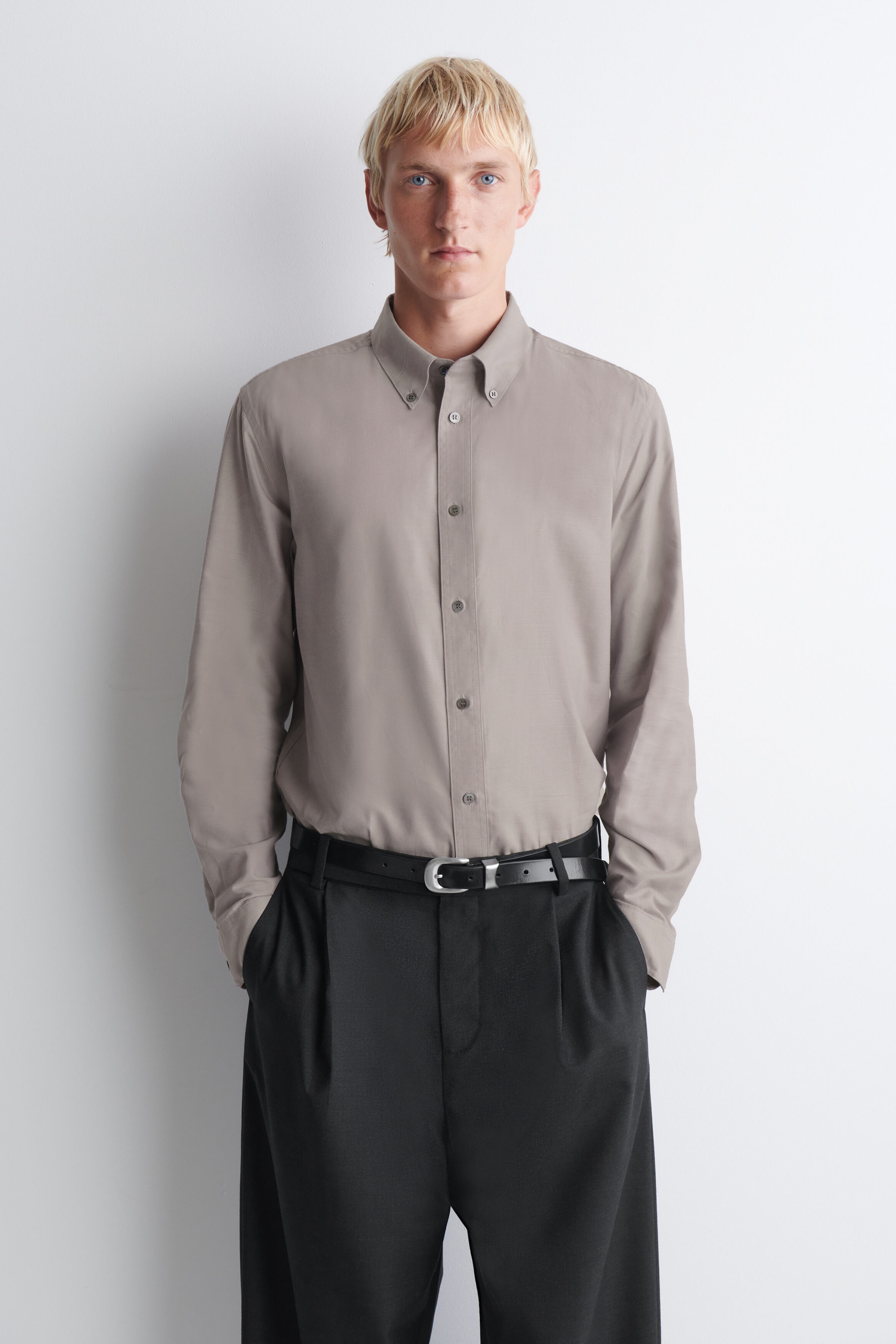 A button-down collar brushed Oxford shirt in a muted taupe shade is worn facing front, tucked into black pleated trousers with a black belt. The shirt features a classic button-down collar, a tonal button placket, and long sleeves.