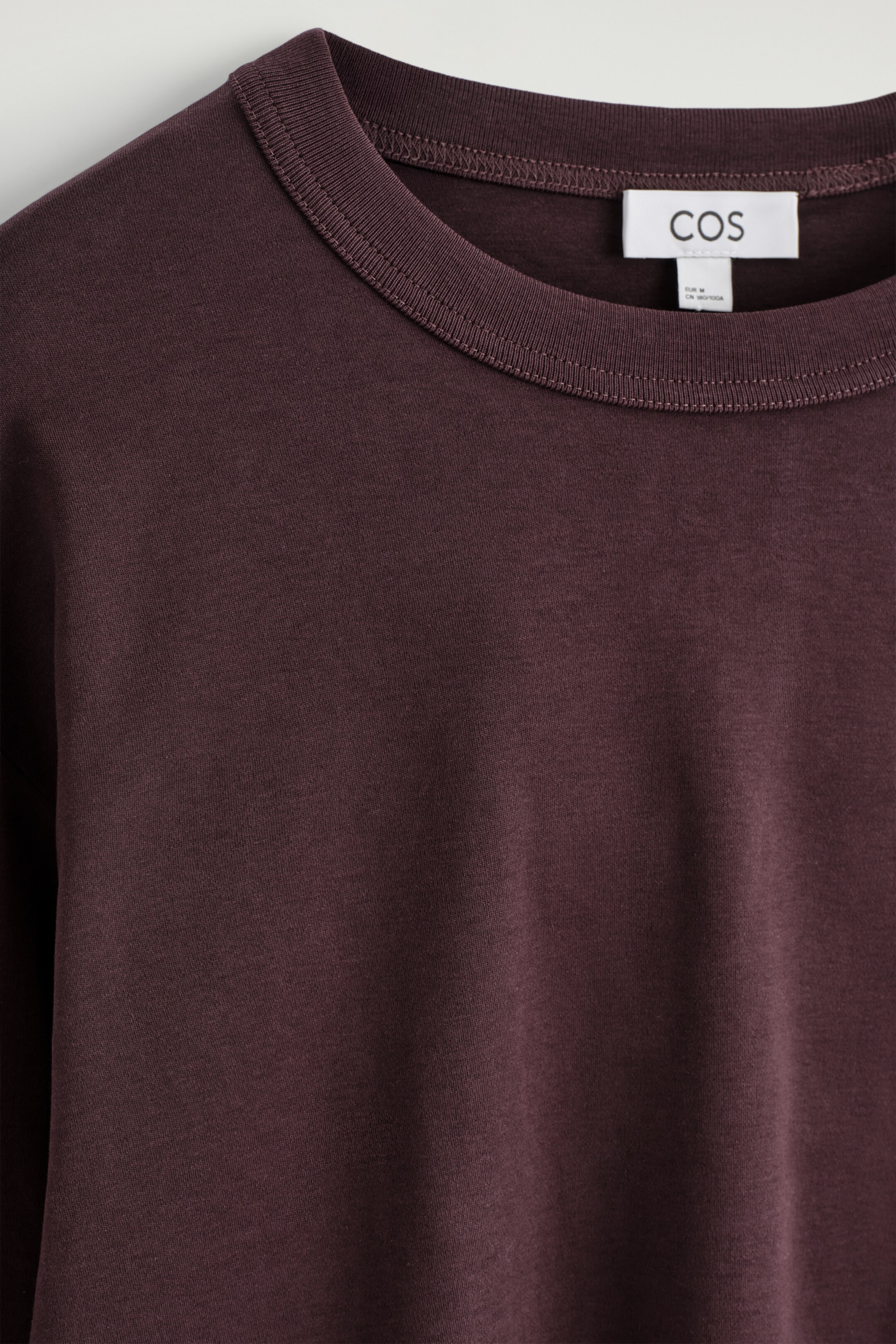 Visualizza immagine più grande: Una vista ravvicinata FRONTALE mostra una T-shirt di colore marrone-rossastro scuro con un classico girocollo. Il tessuto liscio della maglietta è visibile e un'etichetta del marchio bianca appare sul retro del collo.