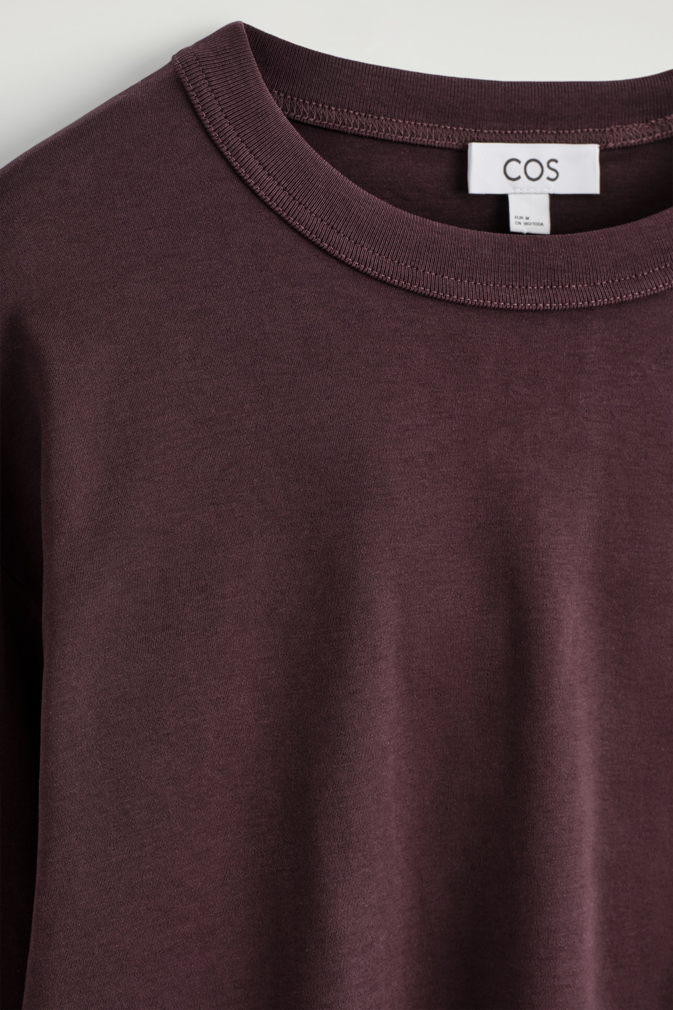 Ver imagen más grande: Una vista FRONTAL en primer plano muestra una camiseta de color marrón rojizo oscuro con un cuello redondo clásico. La tela suave de la camiseta es visible, y una etiqueta de marca blanca aparece en la parte posterior del cuello.