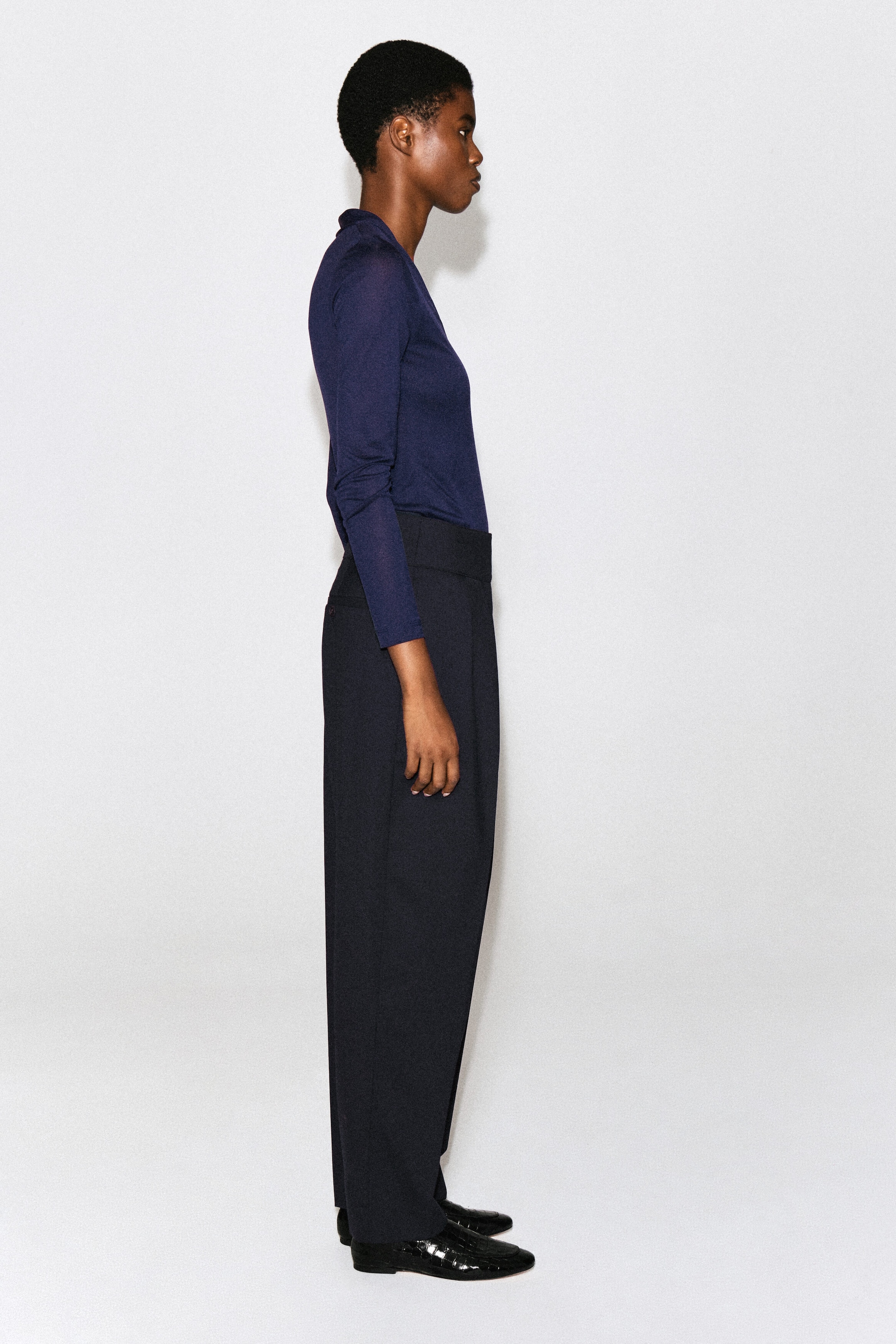 A dark blue, long-sleeved jersey top is being worn by the model, who is standing in a side-profile view. The slim-fit top is tucked into a pair of dark, high-waisted trousers. A dark blue, long-sleeved jersey top is being worn by the model, who is standing in a side-profile view. The slim-fit top is tucked into a pair of dark, high-waisted trousers.