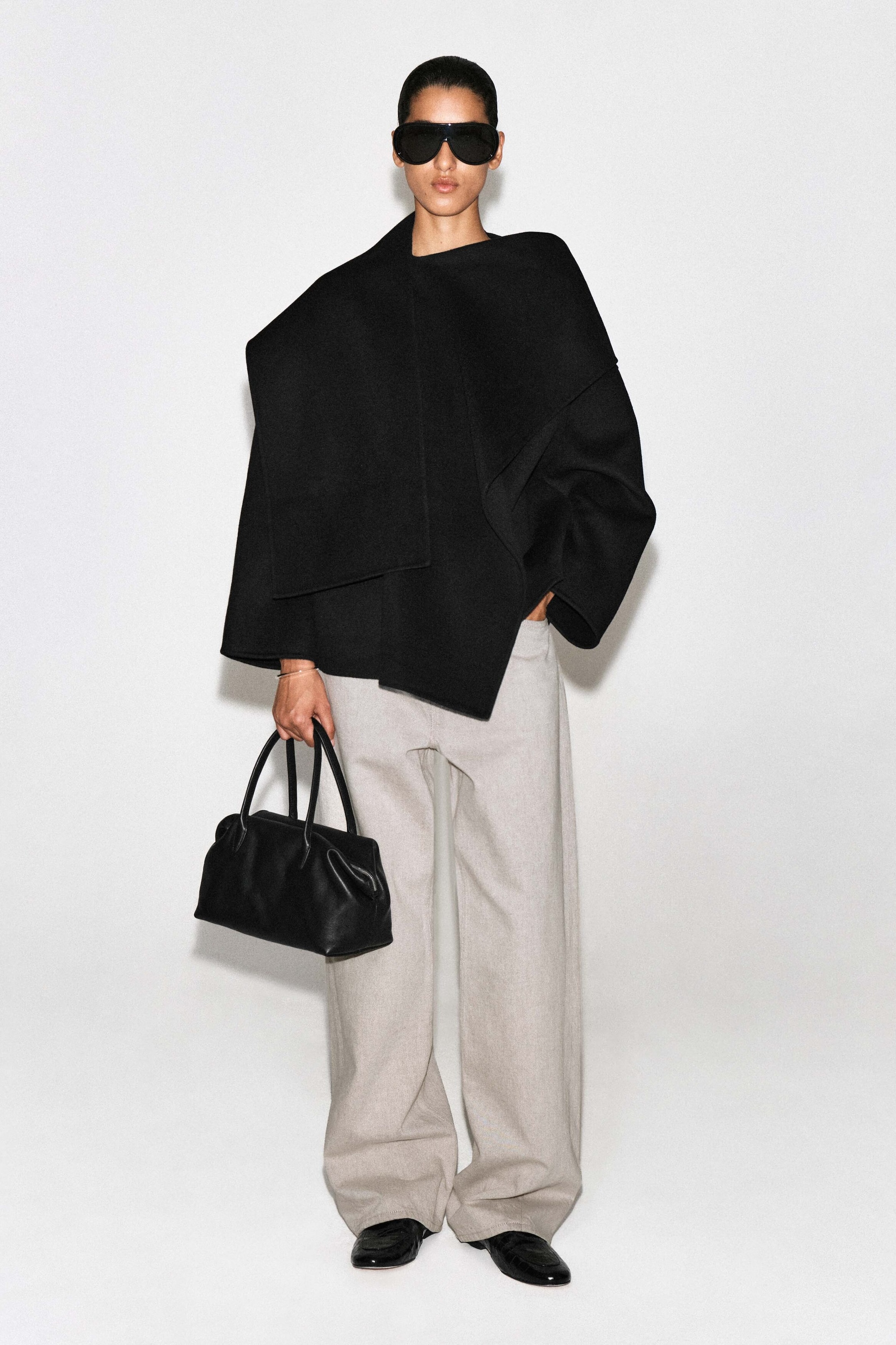 This black double-faced wool-blend scarf jacket is shown from the front, featuring a relaxed silhouette with wide, loose sleeves and an integrated draped scarf collar wrapped over the shoulders. The jacket falls asymmetrically at the hip and is paired with light-colored wide-leg trousers to highlight its drape and structure.