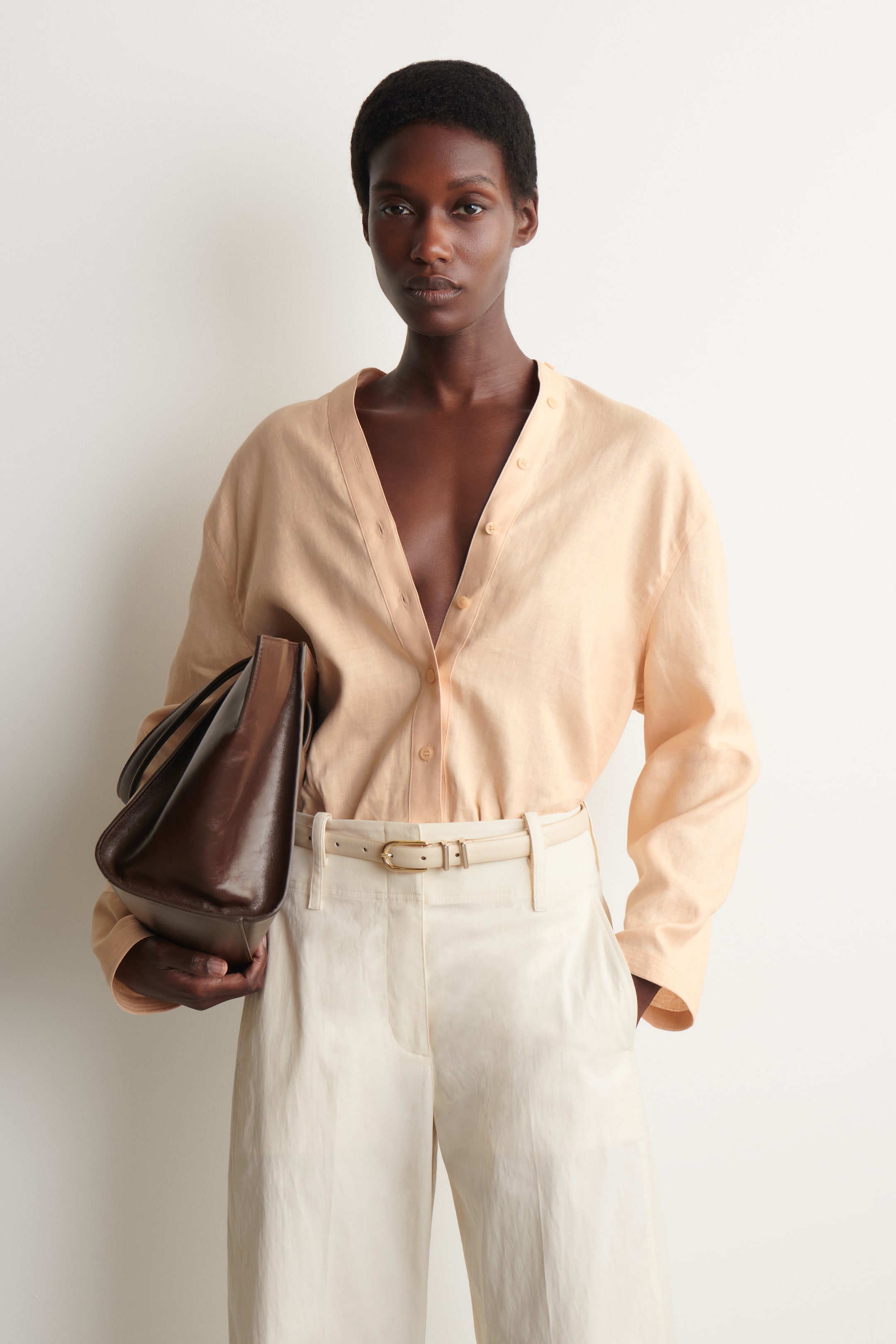 View larger image: The model is wearing a light peach linen shirt, which features a band collar and a V-neck placket with tonal buttons. The shirt is tucked into cream trousers, cinched with a thin belt, and the model is holding a brown leather bag.