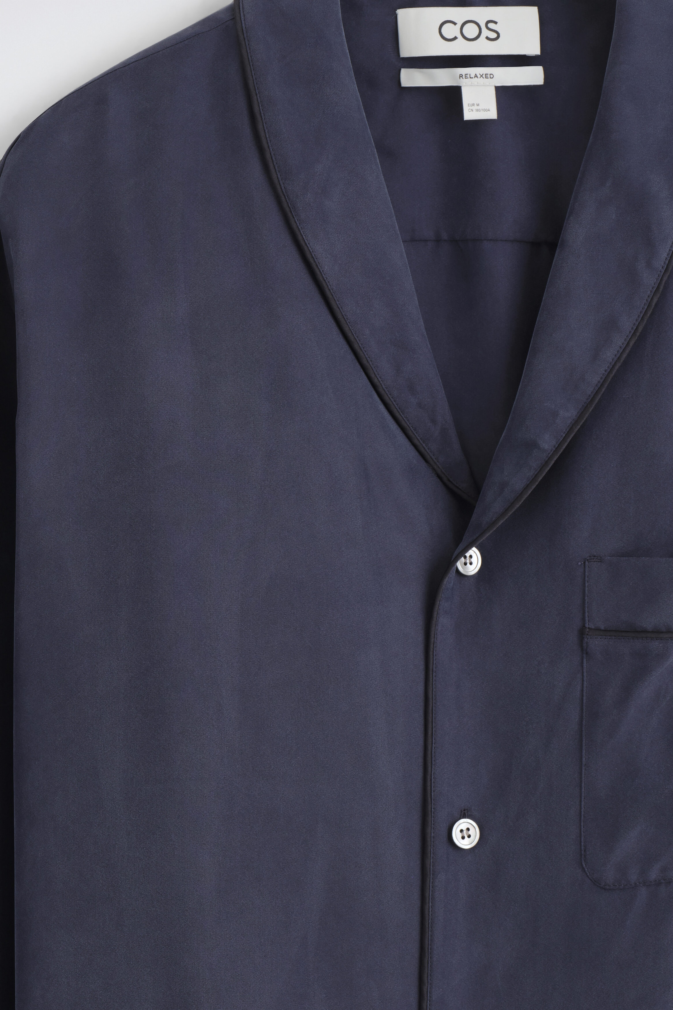 A dark navy silk shirt features a shawl collar with thin contrast piping along the edges and light-coloured mother-of-pearl buttons. Shown from the front, the shirt features a chest patch pocket with matching piped trim. A dark navy silk shirt features a shawl collar with thin contrast piping along the edges and light-coloured mother-of-pearl buttons. Shown from the front, the shirt features a chest patch pocket with matching piped trim.