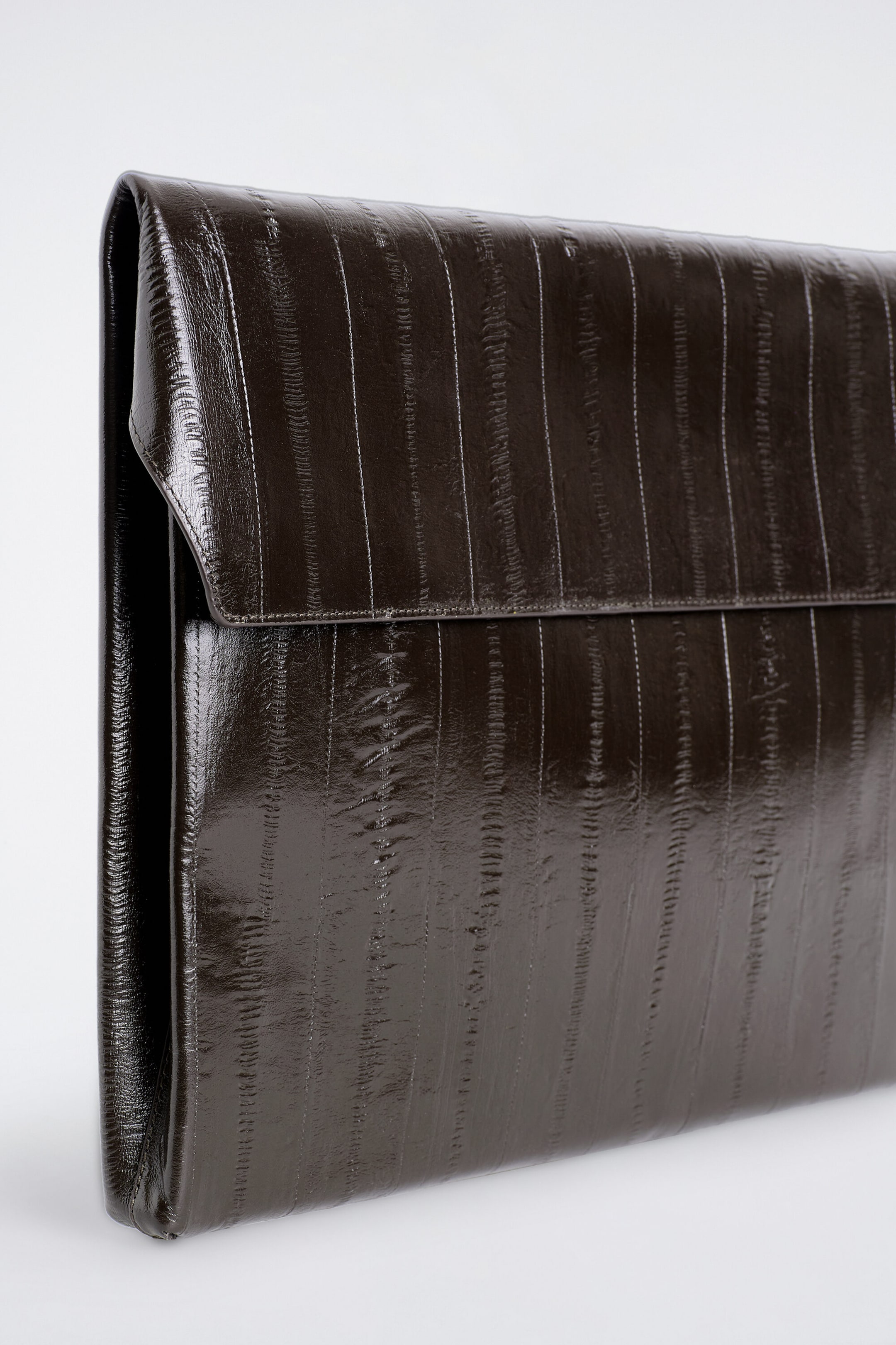 Diese dunkelbraune Leder-Laptophülle im Umschlagstil ist aus einer Dreiviertelansicht dargestellt und hebt ihre strukturierte Silhouette sowie die glänzende, strukturierte Oberfläche mit vertikalen Paneeldetails hervor. Das Design verfügt über einen umklappbaren oberen Klappenverschluss und ein schlankes Profil. Diese dunkelbraune Leder-Laptophülle im Umschlagstil ist aus einer Dreiviertelansicht dargestellt und hebt ihre strukturierte Silhouette sowie die glänzende, strukturierte Oberfläche mit vertikalen Paneeldetails hervor. Das Design verfügt über einen umklappbaren oberen Klappenverschluss und ein schlankes Profil.