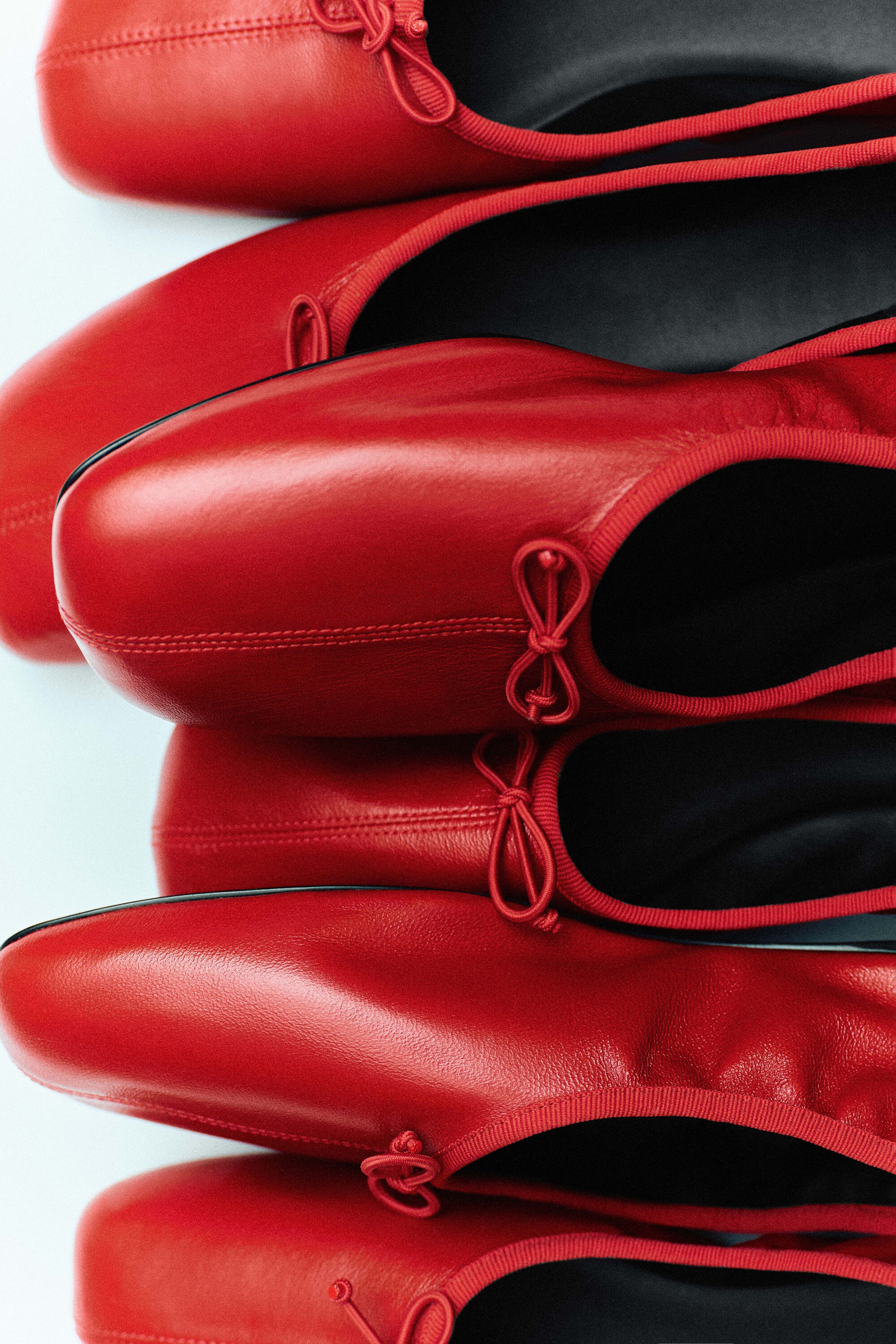 Several red leather ballet flats are overlapping in a stacked vertical arrangement, showcasing their square-toe design with a central top-stitched seam. Each shoe is displaying a red grosgrain edge accented with a small knotted bow and a contrasting black interior.