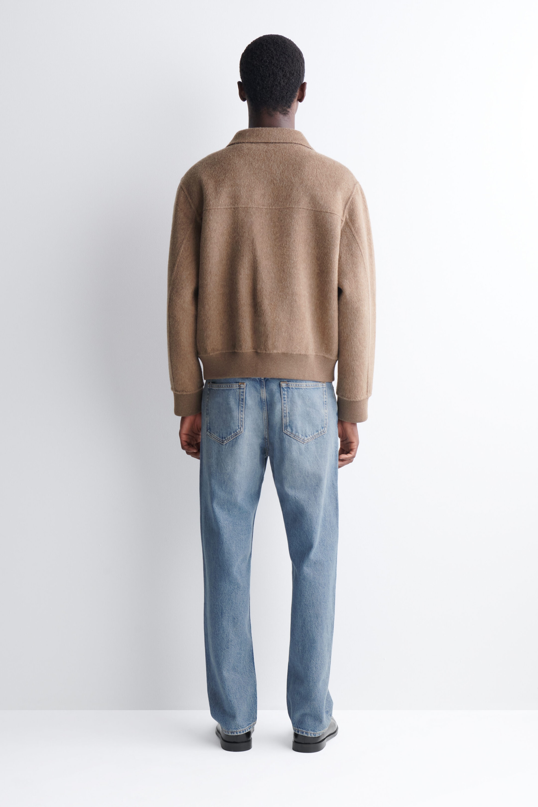 A double-faced blouson jacket in textured beige wool is worn by the model, shown from the back. The jacket features a collar, ribbed cuffs and hem, and a horizontal back yoke, and is styled with light blue jeans. A double-faced blouson jacket in textured beige wool is worn by the model, shown from the back. The jacket features a collar, ribbed cuffs and hem, and a horizontal back yoke, and is styled with light blue jeans.