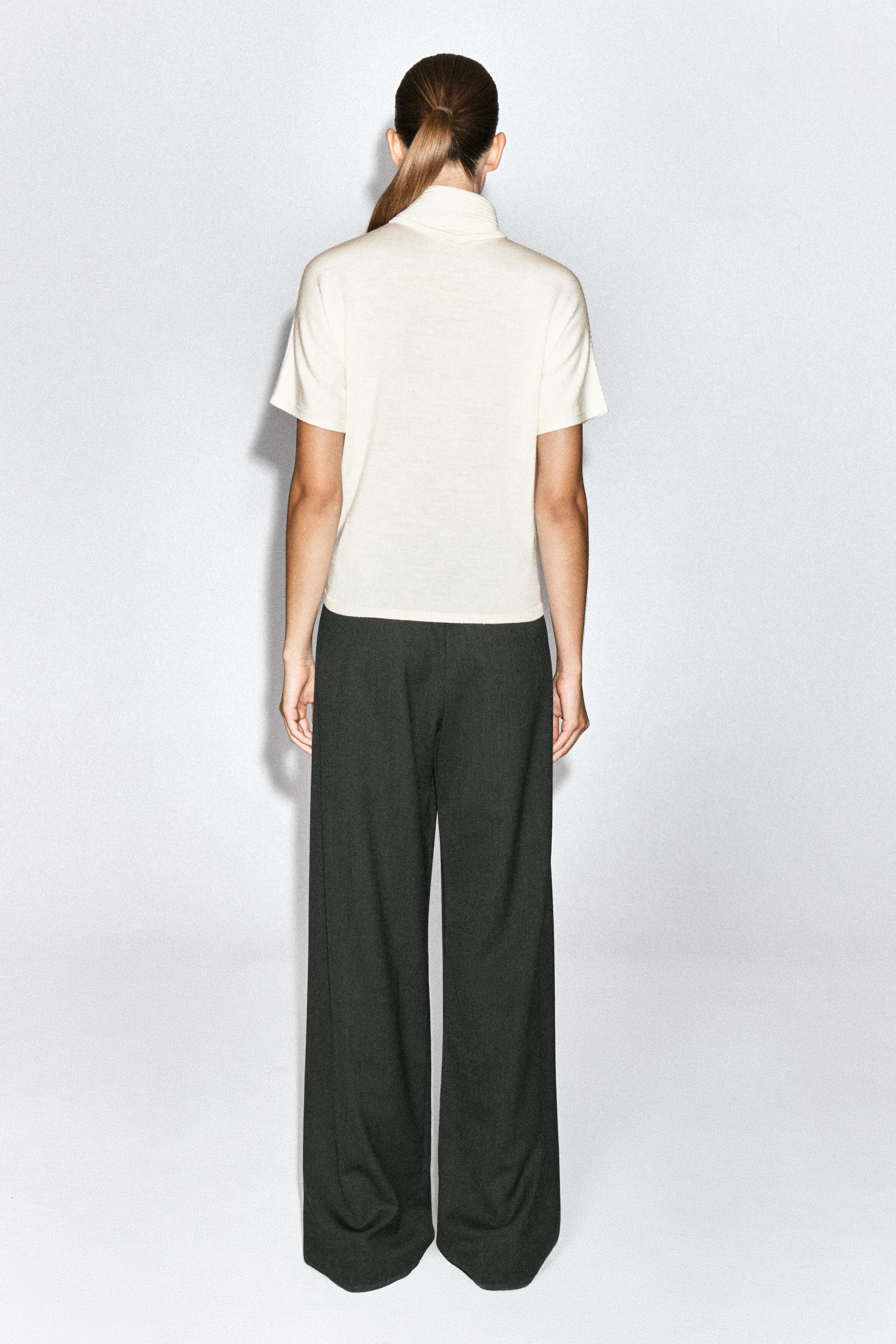 The cream scarf-detail merino wool-blend T-shirt is worn by the model, shown from the back. The short-sleeve top features a high, draped neckline and is styled with dark, wide-leg trousers.