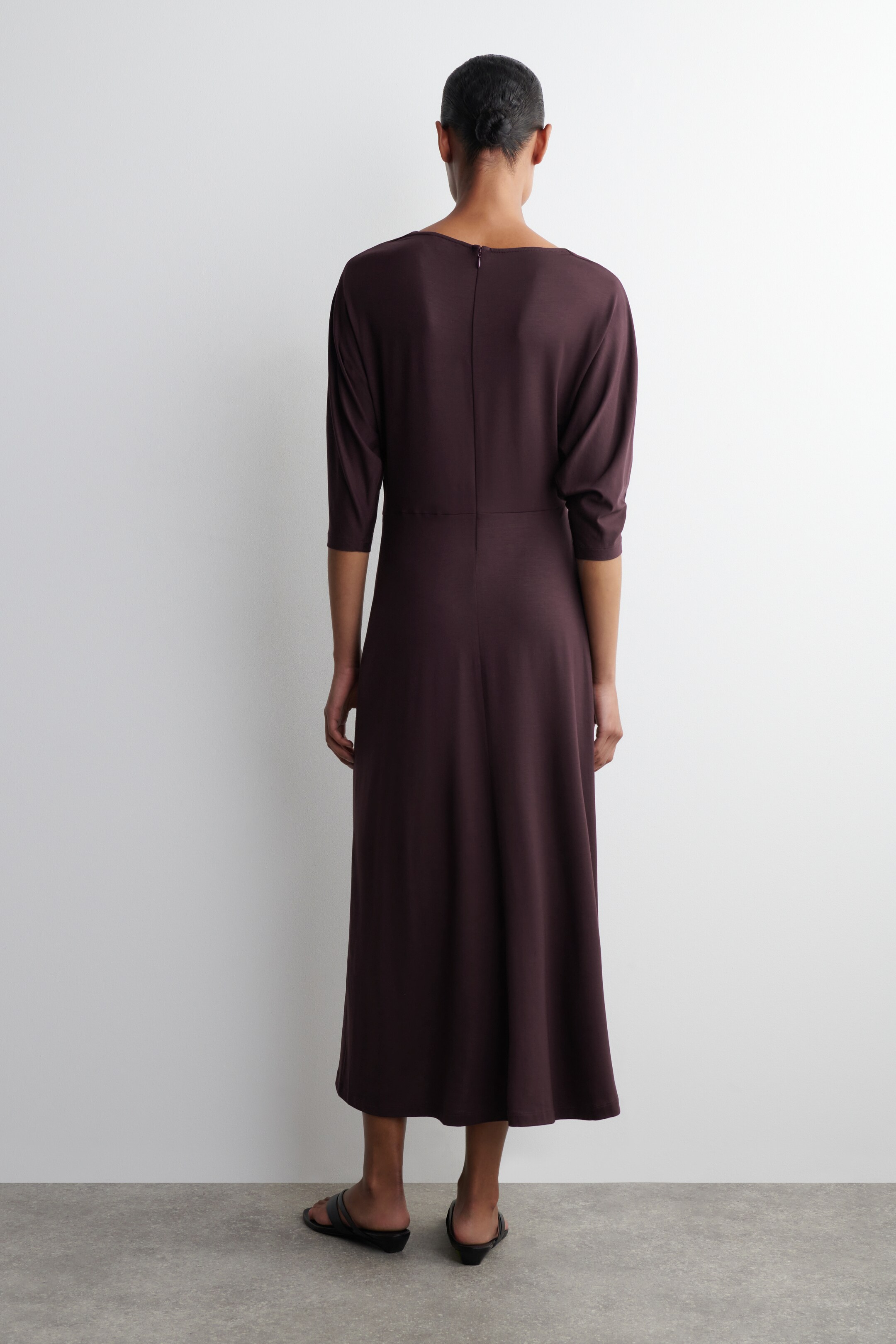 Agrandir l'image: Une robe midi prune foncée est présentée de dos, avec des manches trois-quarts, un large décolleté et une fermeture éclair visible descendant le long du dos. La robe est cintrée à la taille naturelle, et sa jupe fluide s'étend jusqu'à une longueur midi.