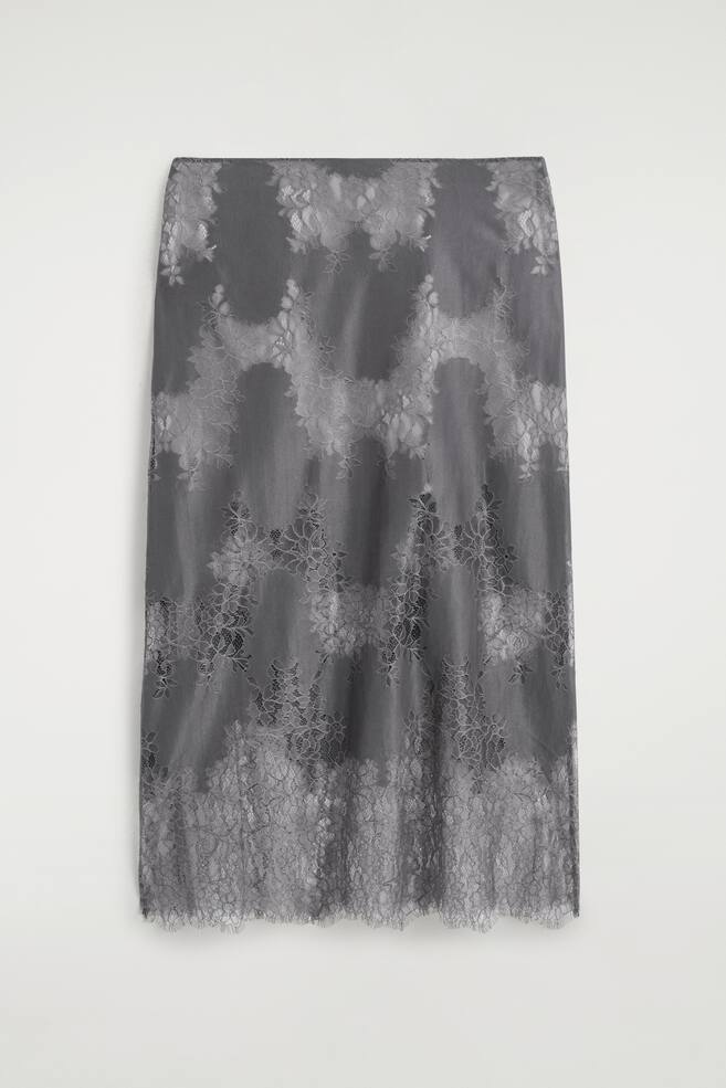 A grey lace-overlay midi skirt is shown from the front, featuring a semi-sheer floral lace overlay draped over a shorter, solid grey lining. The skirt has a straight midi silhouette with a scalloped, eyelash-trimmed lace hem. - 1