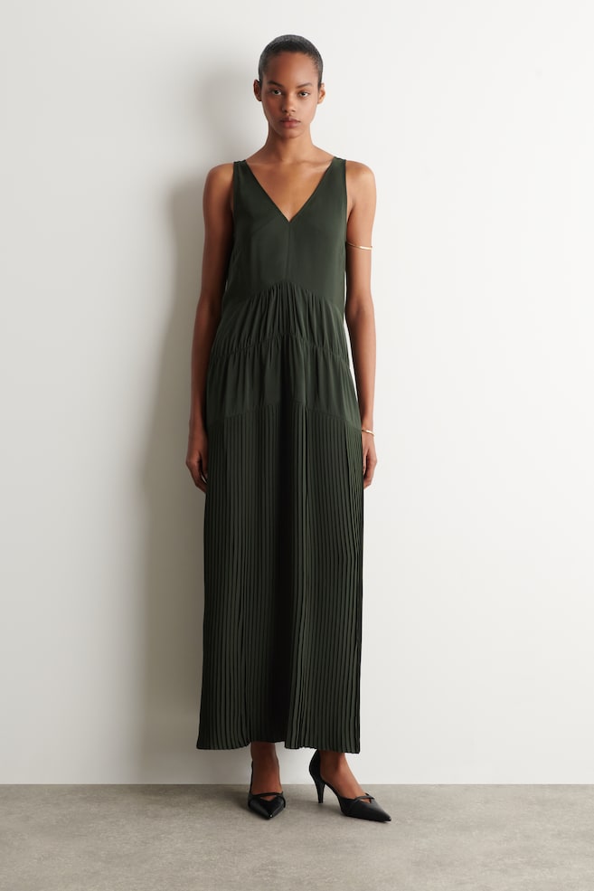 La modella indossa un abito lungo senza maniche verde scuro con scollo a V, caratterizzato da un corpetto liscio, pannelli raccolti a balze e una gonna completamente plissettata. Indossa anche delle scarpe con tacco basso a punta nera. - 1
