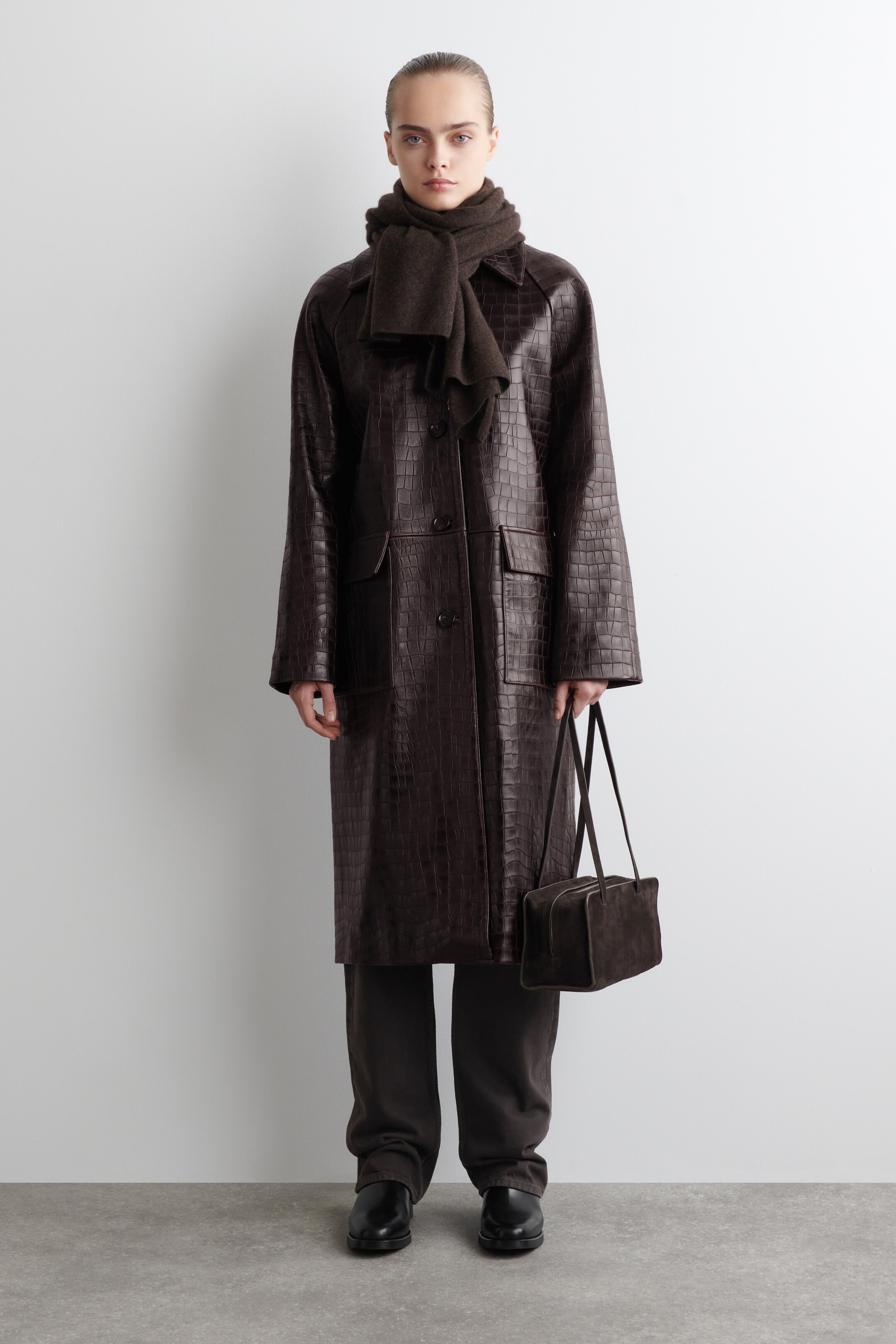 Agrandir l'image: Un manteau long gaufré de couleur marron foncé, avec un motif croco, est orienté vers l'avant, présentant une fermeture à boutons sur le devant et deux grandes poches à rabat sur le devant. Une écharpe assortie de couleur marron foncé est enroulée autour du cou, couvrant partiellement le col du manteau.