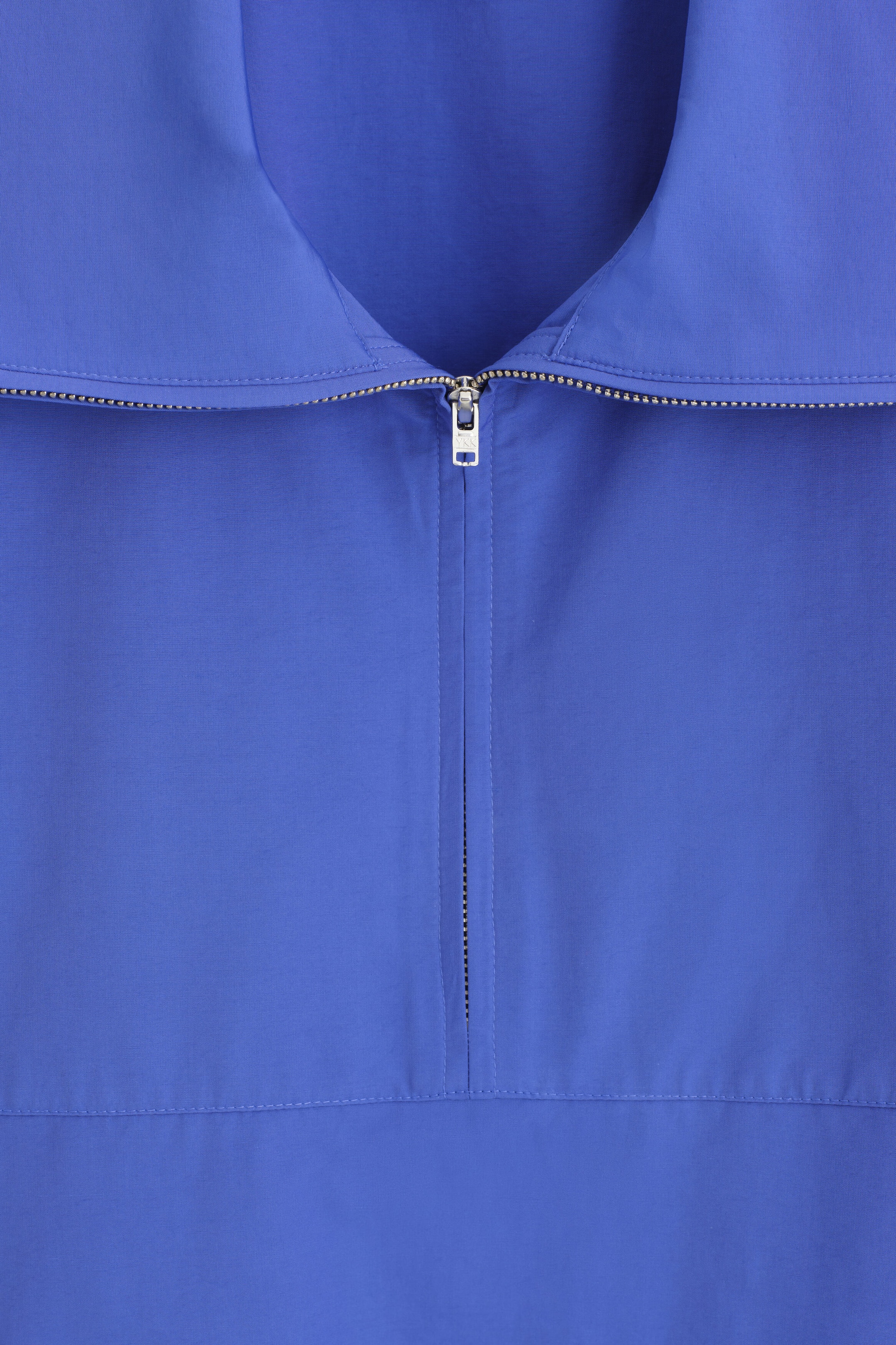 A close-up front view of a blue garment showcasing a wide collar, a silver-tone half-zip fastening, and fine topstitched seams. A close-up front view of a blue garment showcasing a wide collar, a silver-tone half-zip fastening, and fine topstitched seams.