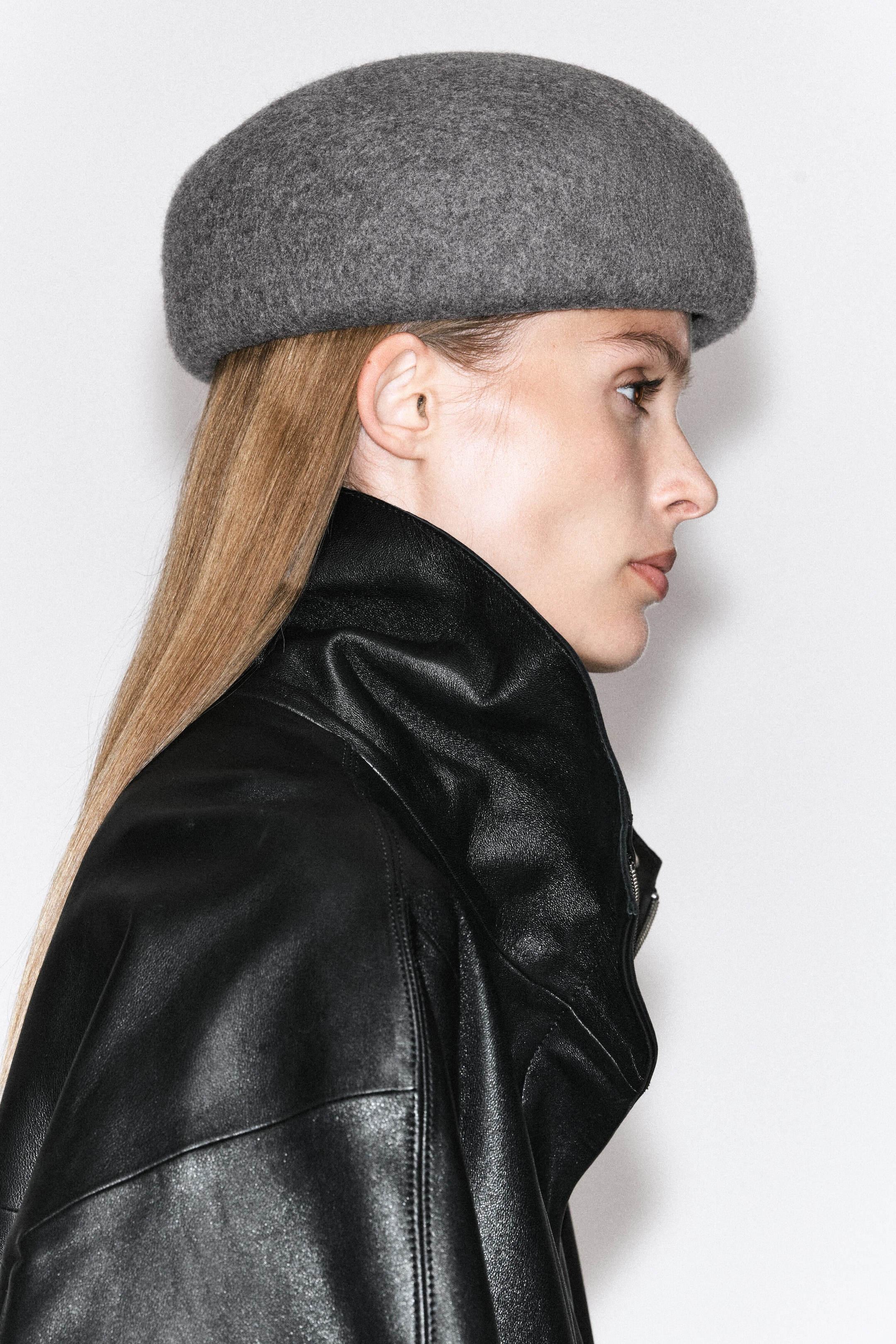 A sculpted wool beret in a textured grey mélange is being worn by the model, shown in profile. The structured, rounded hat sits low on the head and is styled with a high-collared black leather jacket. A sculpted wool beret in a textured grey mélange is being worn by the model, shown in profile. The structured, rounded hat sits low on the head and is styled with a high-collared black leather jacket.