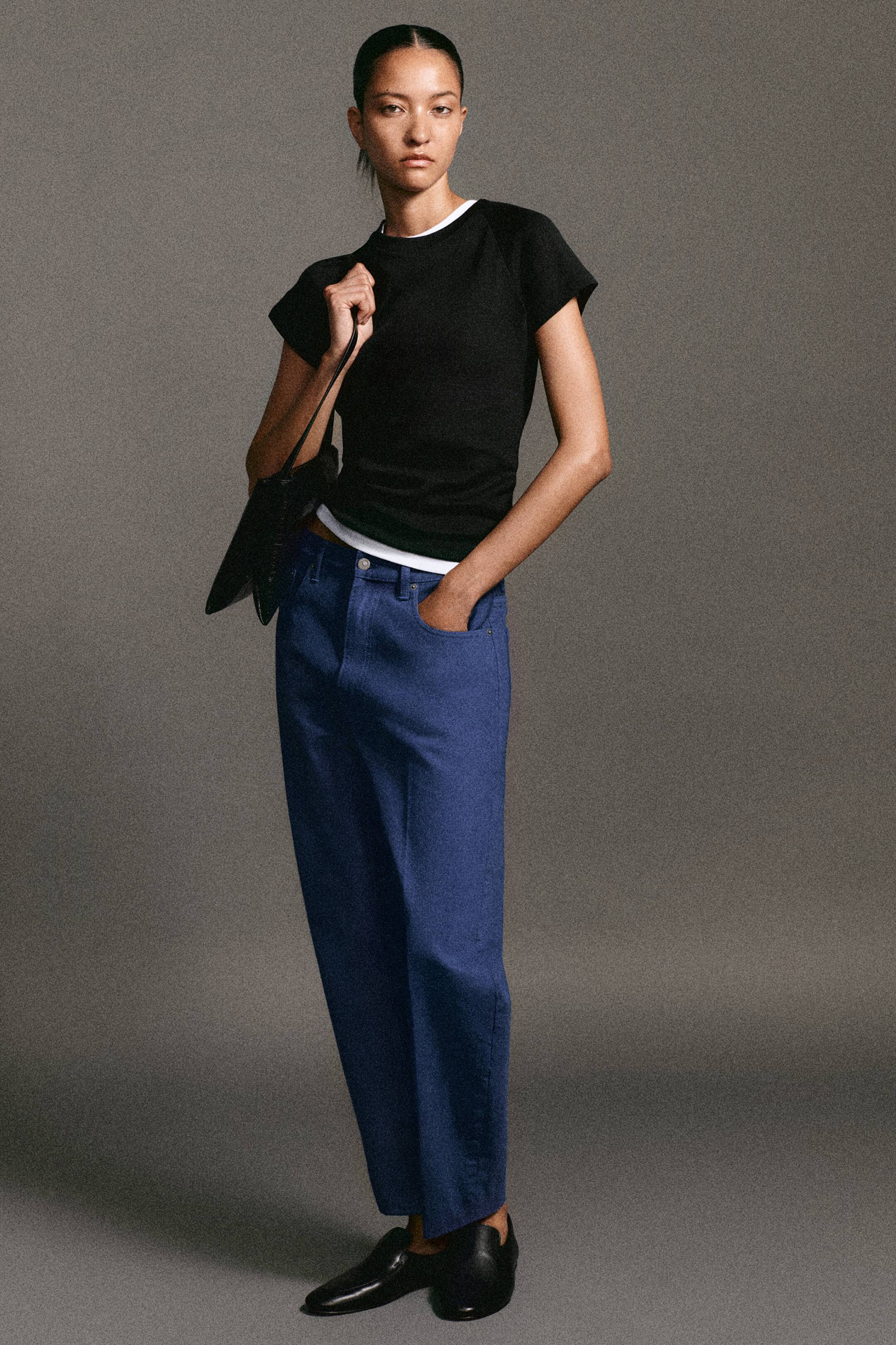 Vezi imaginea mai mare: Văzut din față, un model poartă tricoul raglan slim din bumbac negru, purtat în straturi peste un tricou alb, vizibil la guler și la tiv. Ea asortează partea de sus cu blugii conici Umbra albaștri, care au talie înaltă, croială relaxată și pliuri structurate pe partea din față a pantalonului.