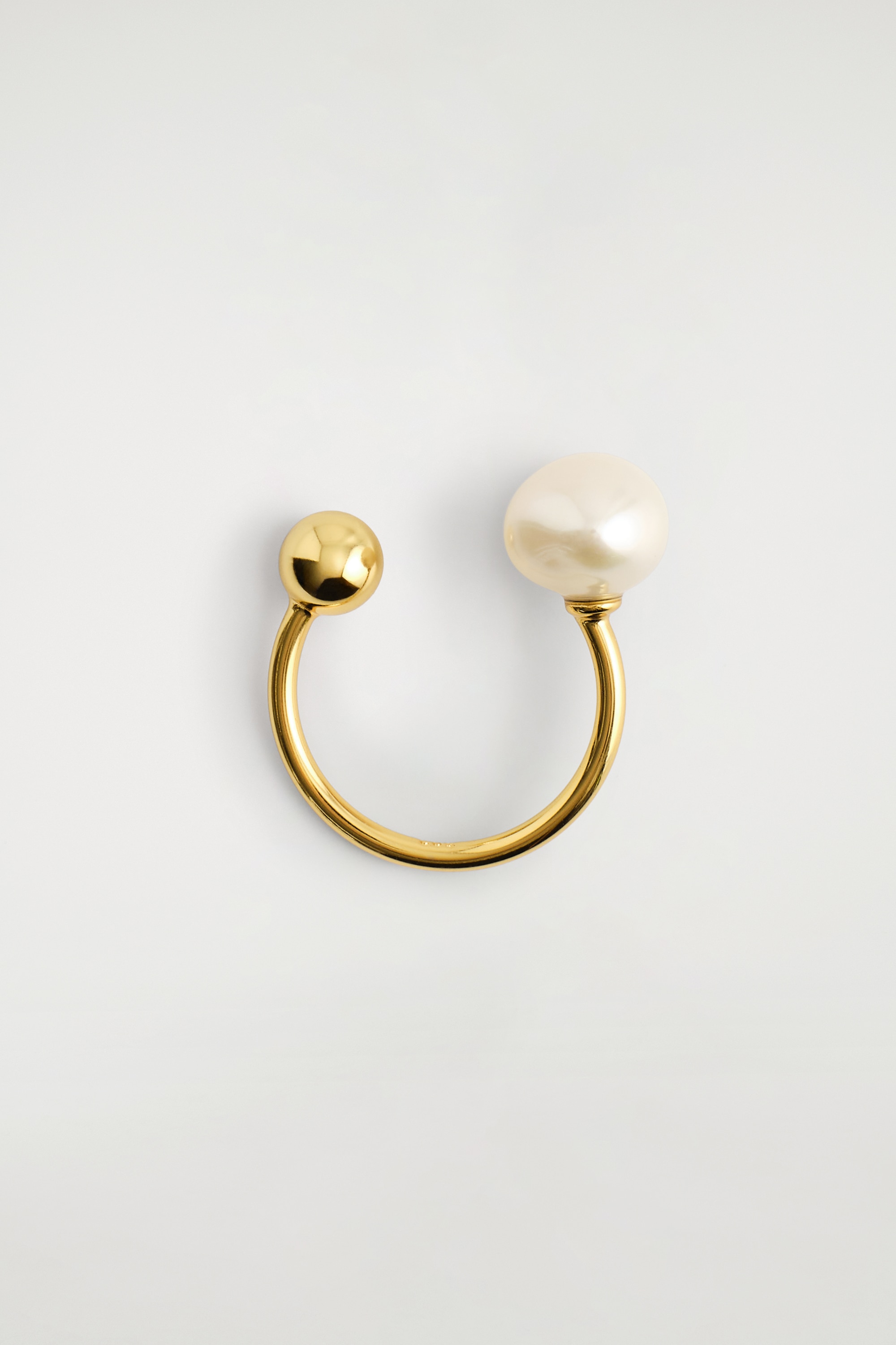 Een open ring in goudtint met een witte zoetwaterparel en een kleine gouden bol wordt gedragen aan de vinger van het model. De hand rust tegen een witte denim broek, terwijl de mouw van een zwart leren jack net boven de pols eindigt.