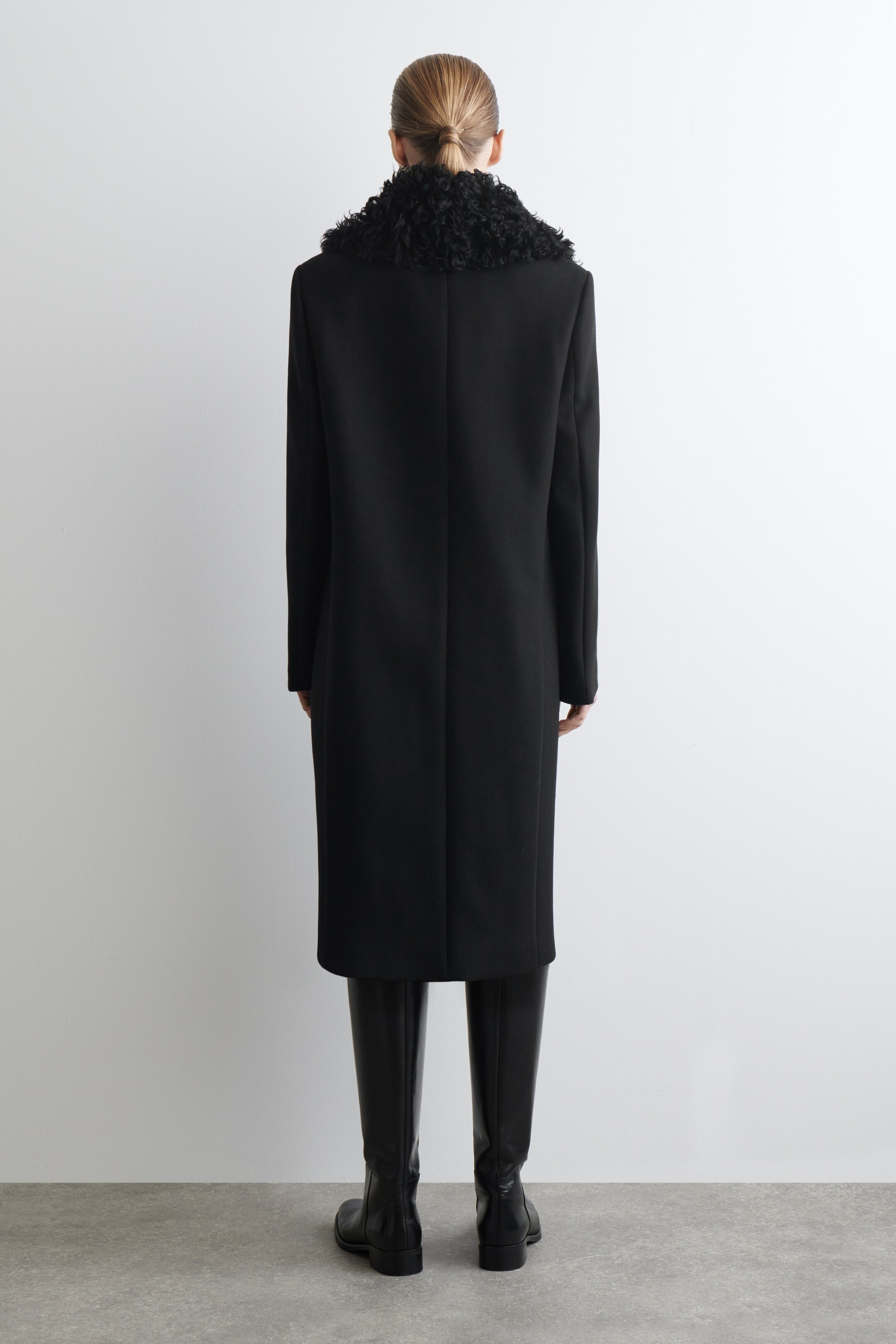 View larger image: A black coat is being presented from the back. It is featuring an oversized, curly black shearling collar and a tailored, straight silhouette that extends to knee-length, with a vertical seam running down the center back.