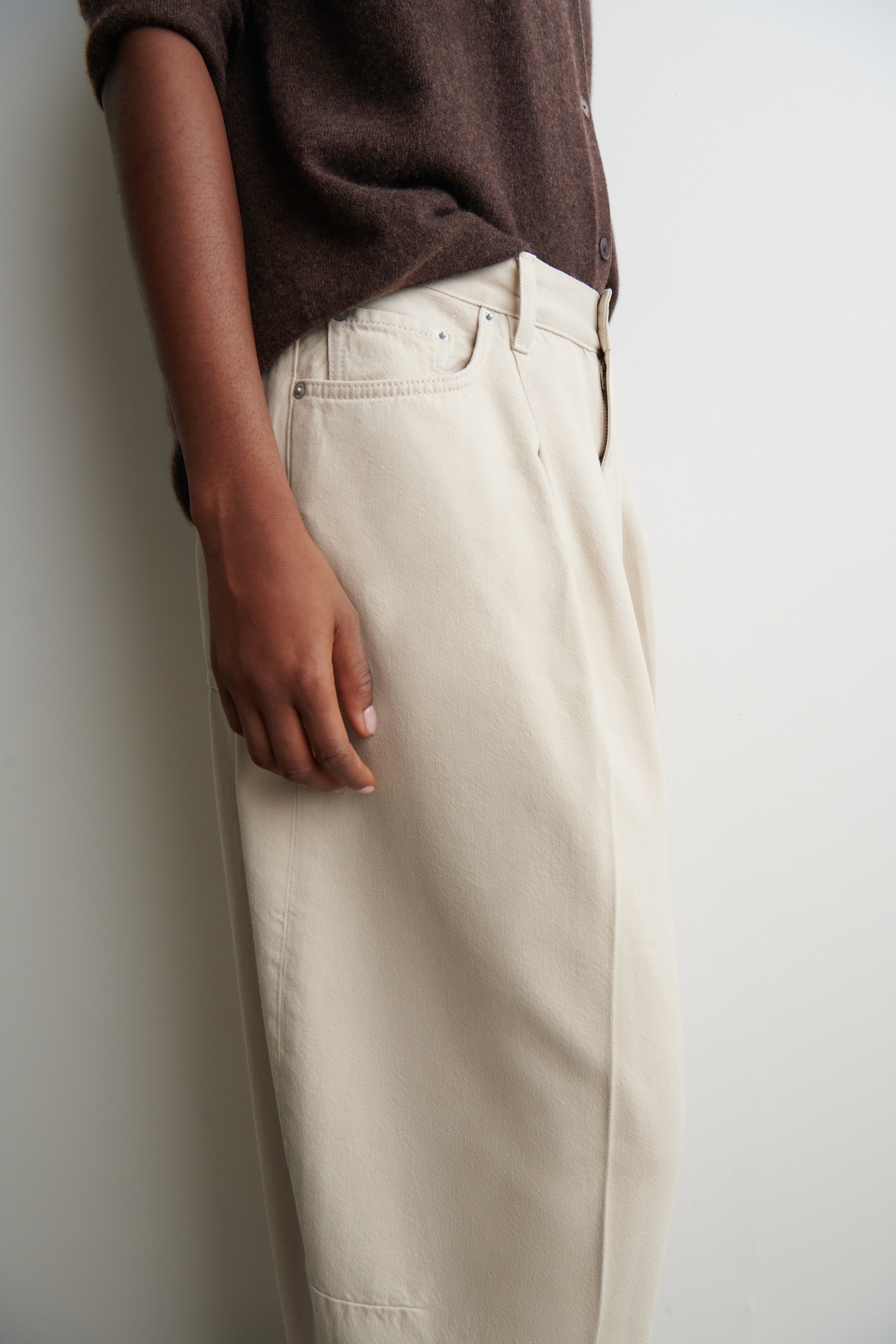 View larger image: Beige trousers are being shown from a side angle, revealing the upper left hip and leg. They are featuring front slash pockets with rivets, a small coin pocket, and a visible vertical seam running down the side of the leg. A dark brown cardigan is partially tucked into the waistband.