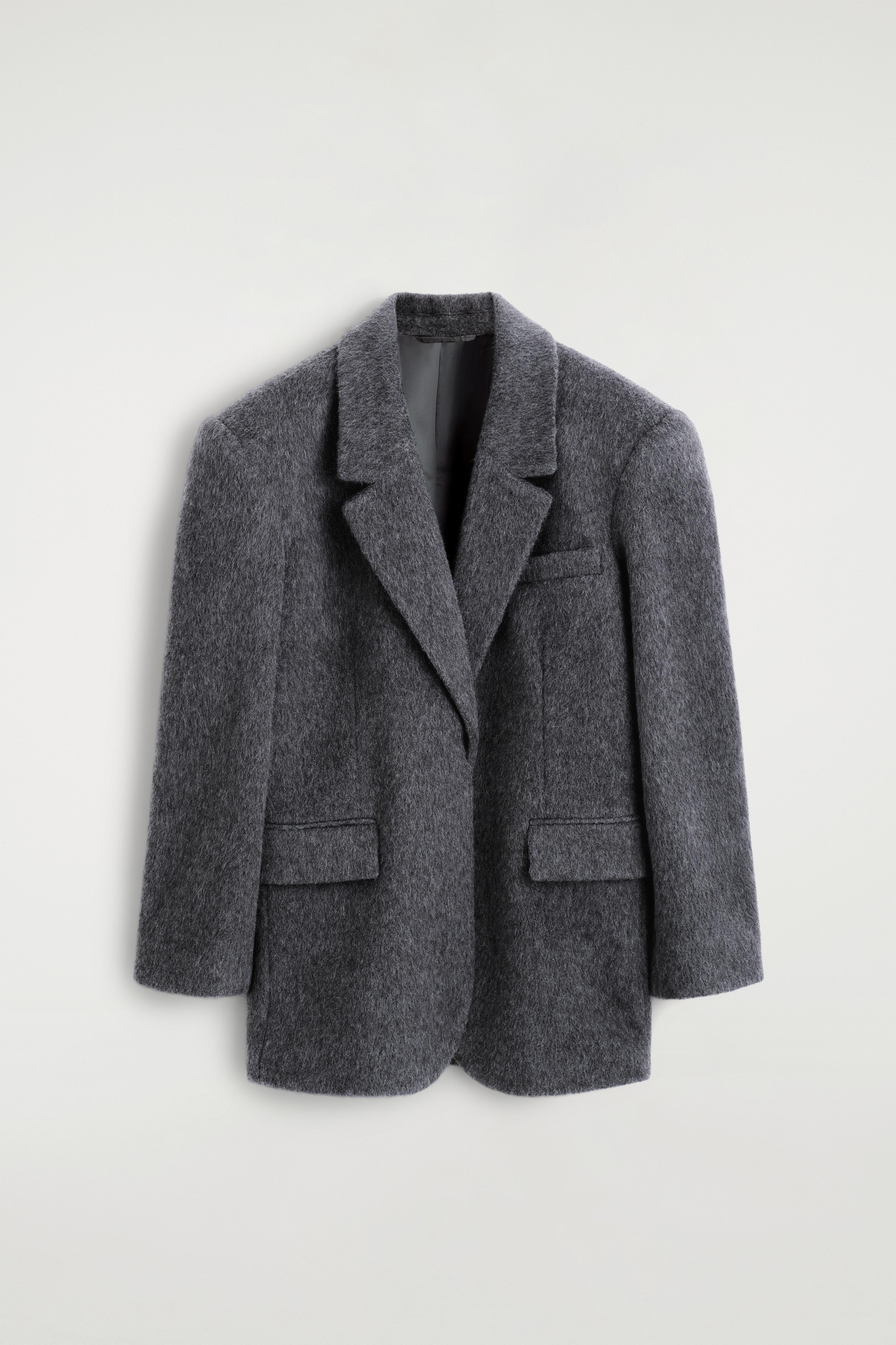 En kolgrå, texturerad blazer i ullblandning med slag och en avslappnad passform bärs öppen av modellen. Blazern är stylad över en ljus, randig skjorta med knäppning och blå jeans, med en svart axelväska i läder som bärs på ena sidan.