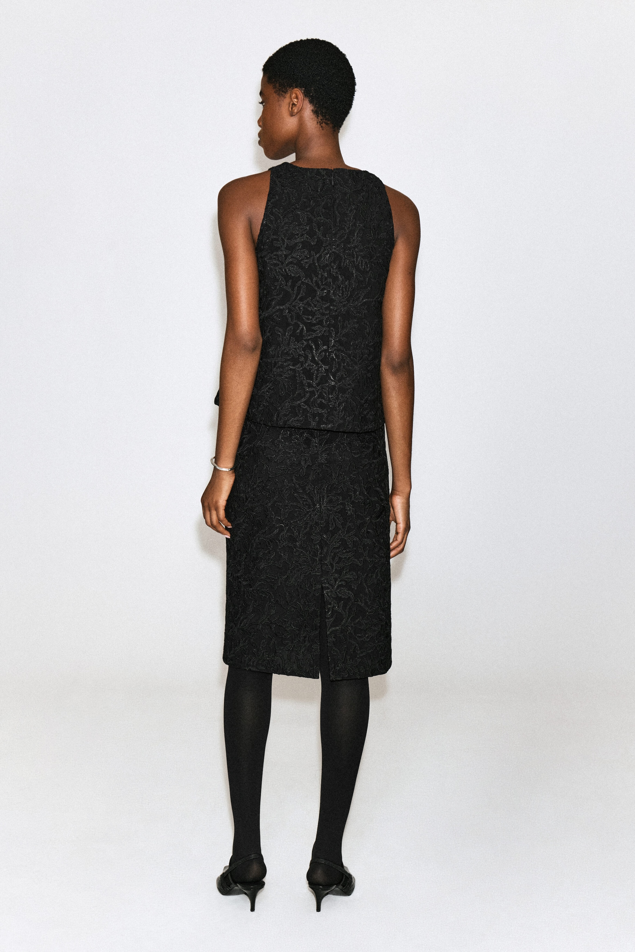 Un haut noir sans manches orné d’une broderie florale texturée est porté par le mannequin, qui est de dos à la caméra. Le haut est assorti à une jupe noire longueur genou, à des collants noirs et à des chaussures noires à petit talon.