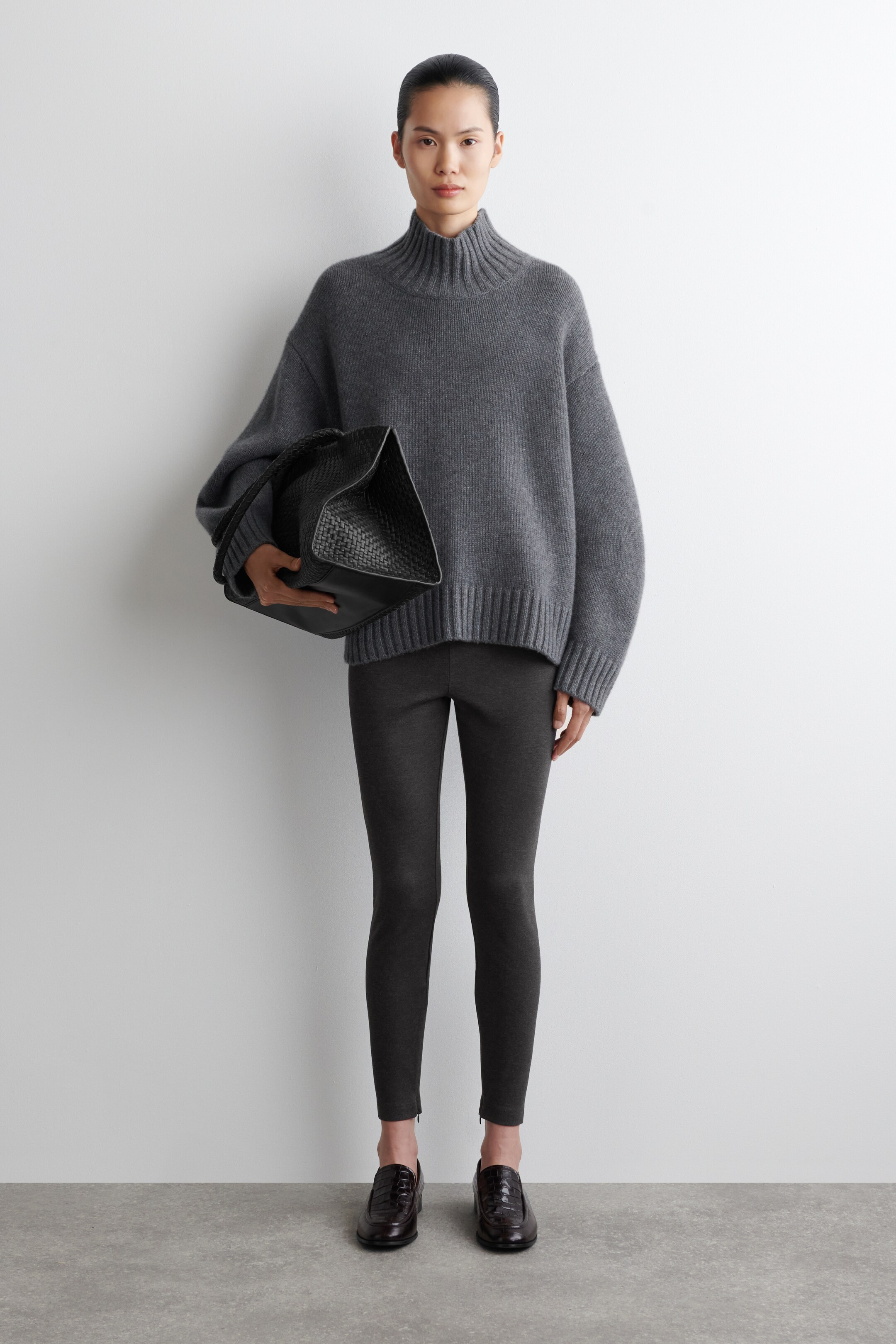Grotere afbeelding bekijken: Een grijze, oversized coltrui met een geribde kraag, manchetten en zoom wordt aan de voorkant getoond, gedragen met donkergrijze slim-fit enkelbroeken met zichtbare enkelritsen. Een zwarte geweven tote bag wordt vastgehouden en er worden donkerbruine loafers met textuur gedragen.