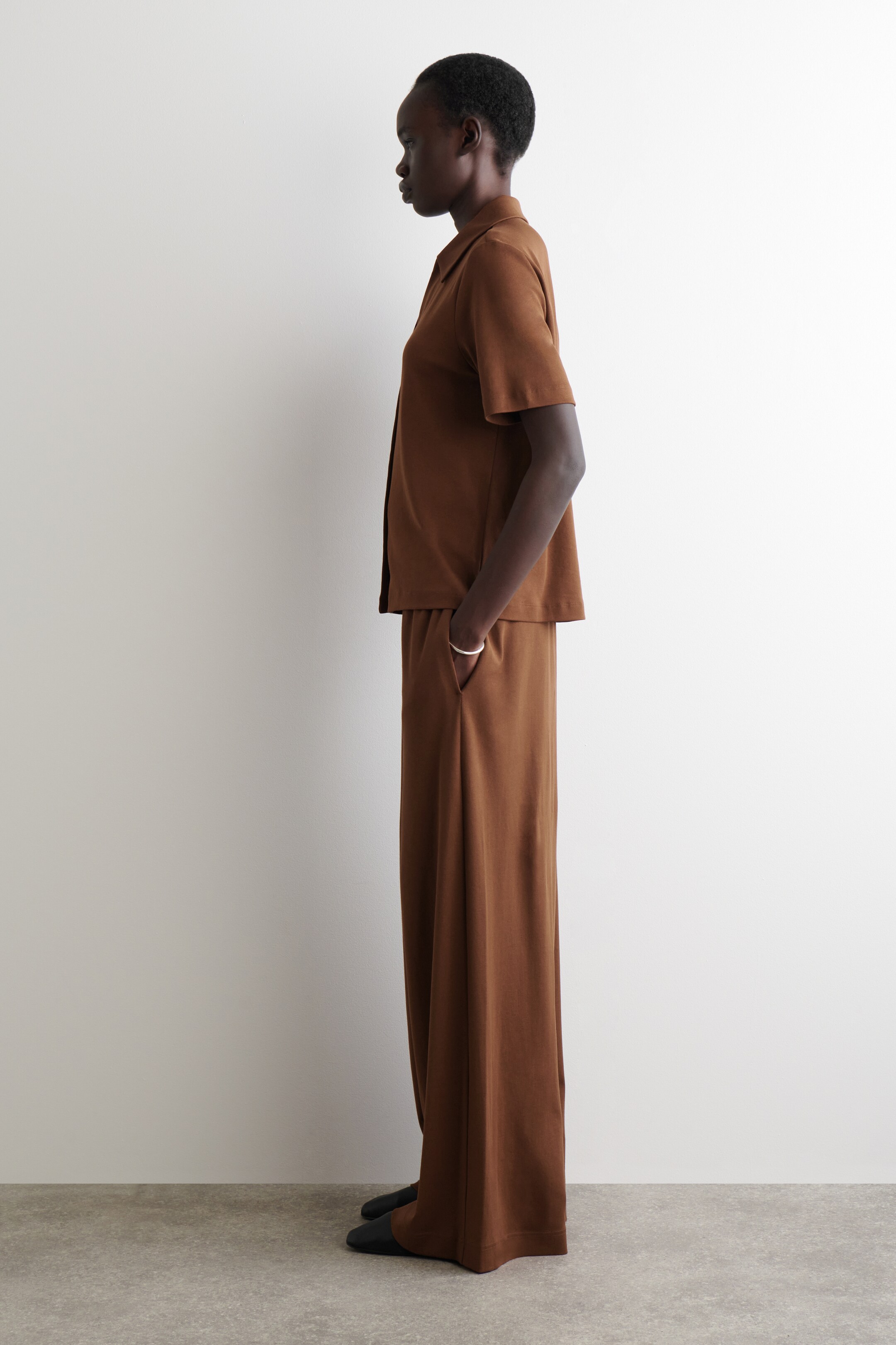 Agrandir l'image: Un ensemble deux pièces marron, composé d'un haut à manches courtes avec boutons sur le devant et d'un pantalon à jambes larges, est présenté de profil. Le haut présente un col pointu et une silhouette décontractée, tandis que le pantalon assorti a une coupe fluide avec des poches latérales.