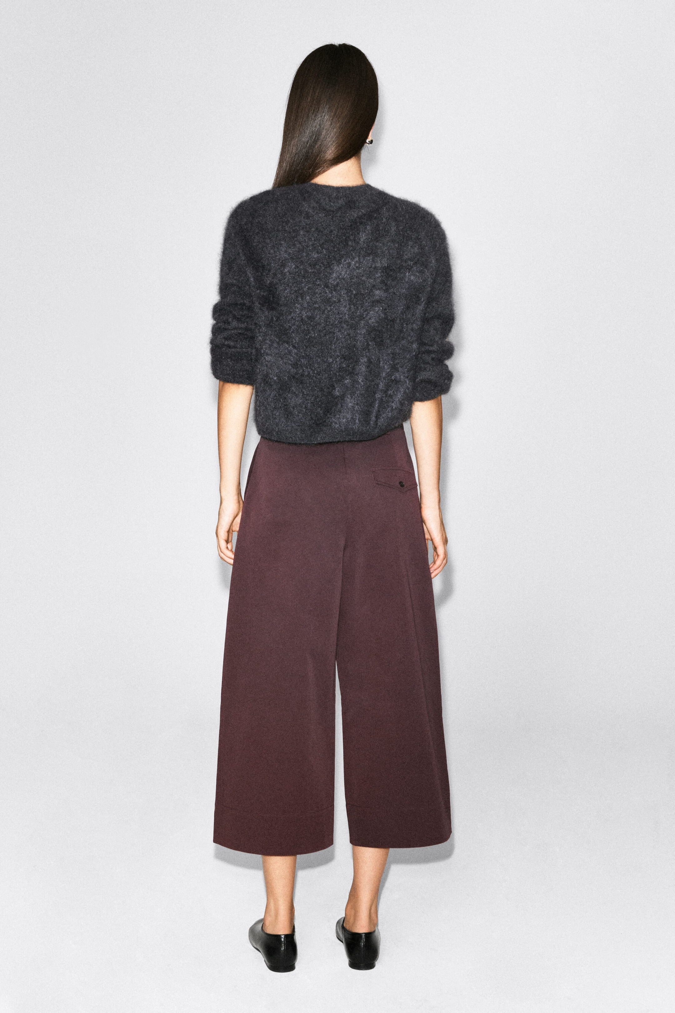 Cotton cropped barrel-leg pants in dark plum are worn by the model, showcasing a wide, curved silhouette and a single buttoned back pocket. The pants are styled with a fuzzy dark gray sweater and black leather flats. Cotton cropped barrel-leg pants in dark plum are worn by the model, showcasing a wide, curved silhouette and a single buttoned back pocket. The pants are styled with a fuzzy dark gray sweater and black leather flats.