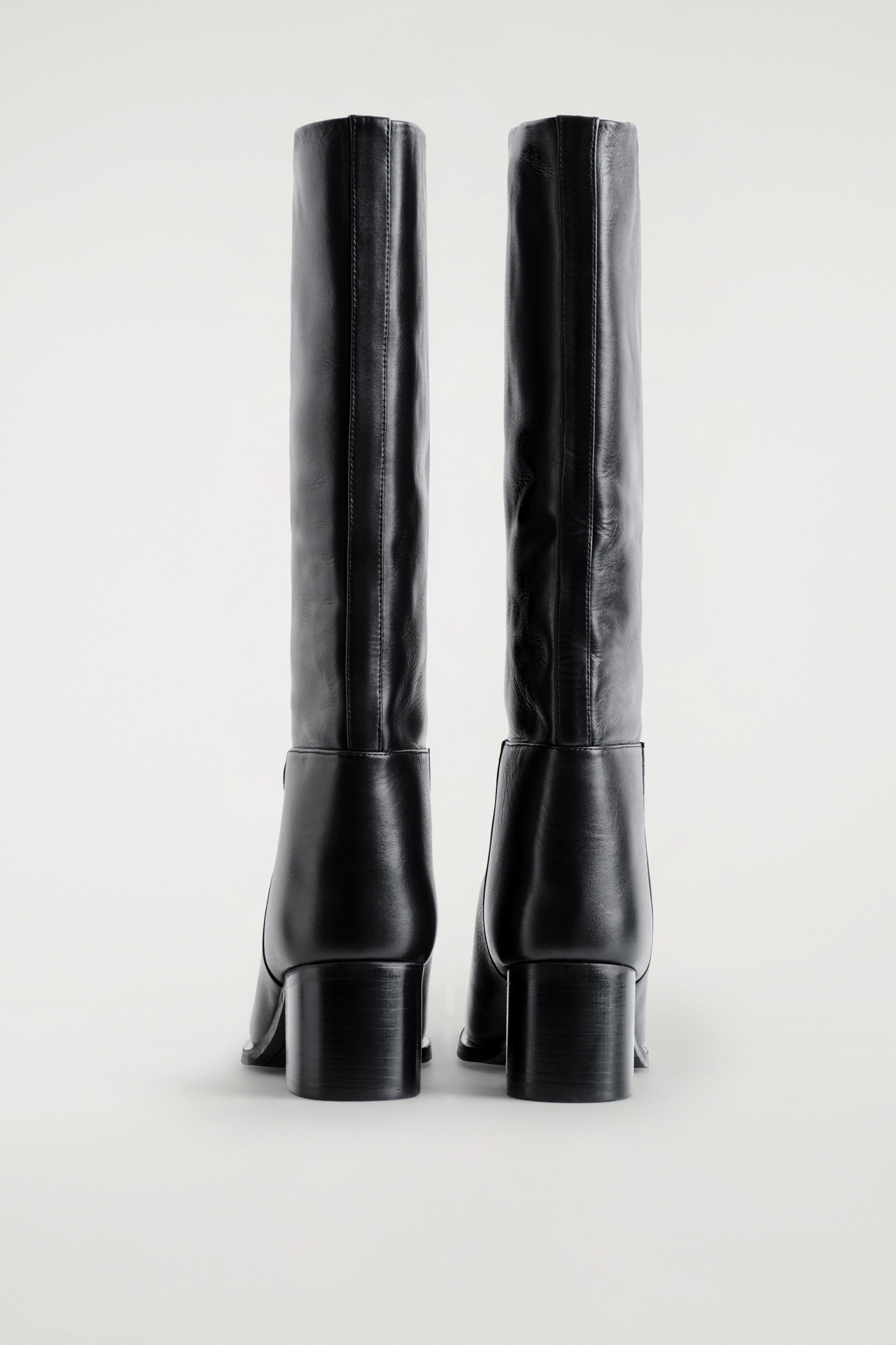 Grösseres Bild ansehen: Schwarze Kniehohe Stiefel werden von hinten gezeigt, mit hohen, geraden Schäften, die eine zentrale vertikale Naht und robuste Blockabsätze aufweisen.